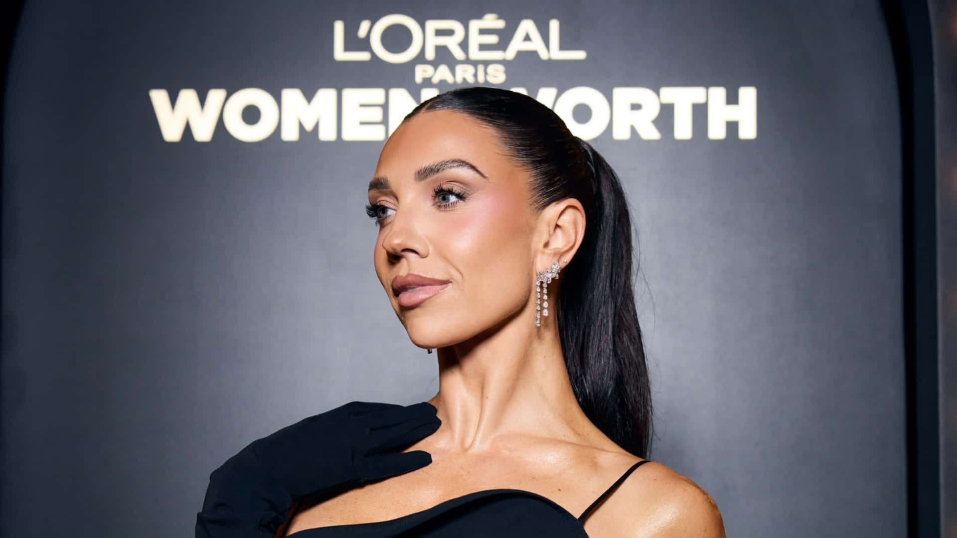  壁紙 — 一位優雅的女人穿著黑色連衣裙和手套，在帶有L'Oréal Paris Women Worth品牌的深色背景下自信地擺姿勢。