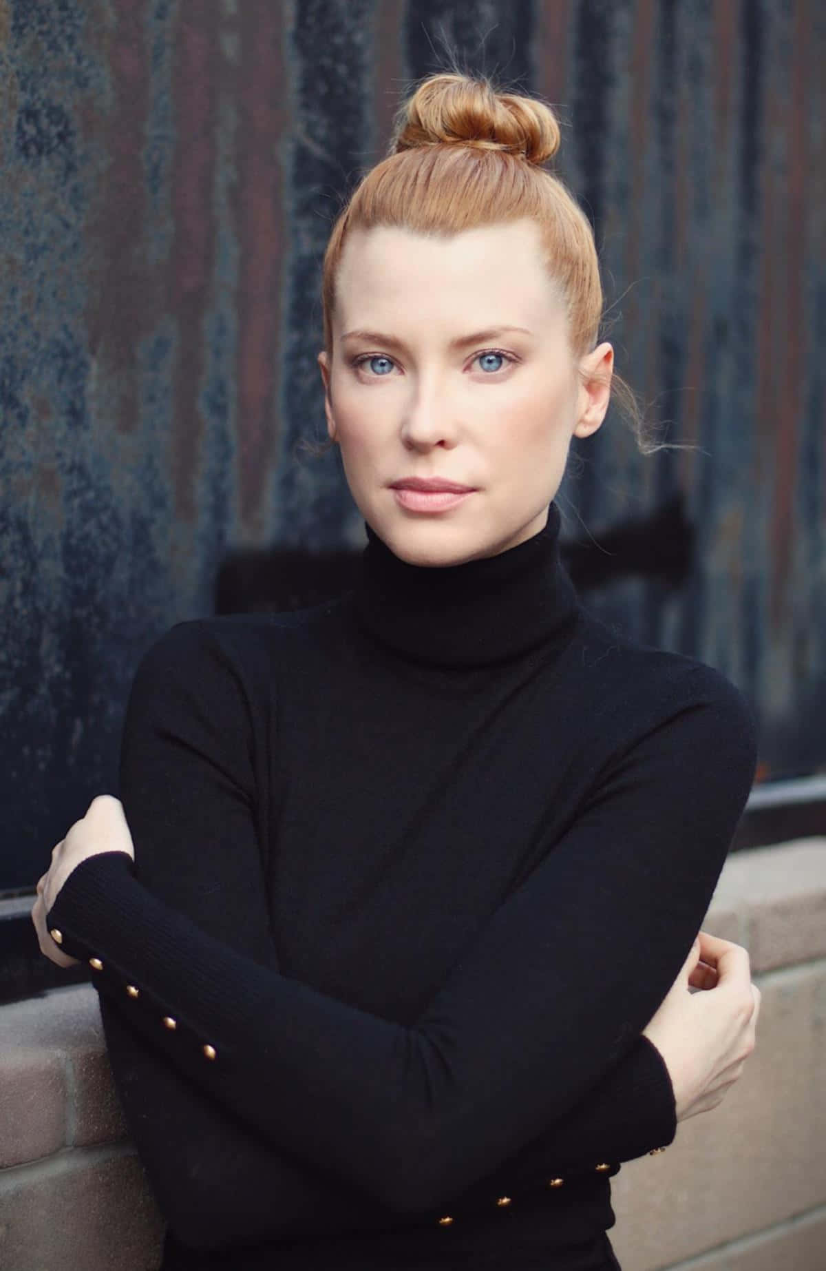 Emma Booth 桌布 壁紙 — 一位紅髮高髻的優雅女士自信地站在風化的金屬牆前，身穿帶有金色鈕扣的黑色高領毛衣。