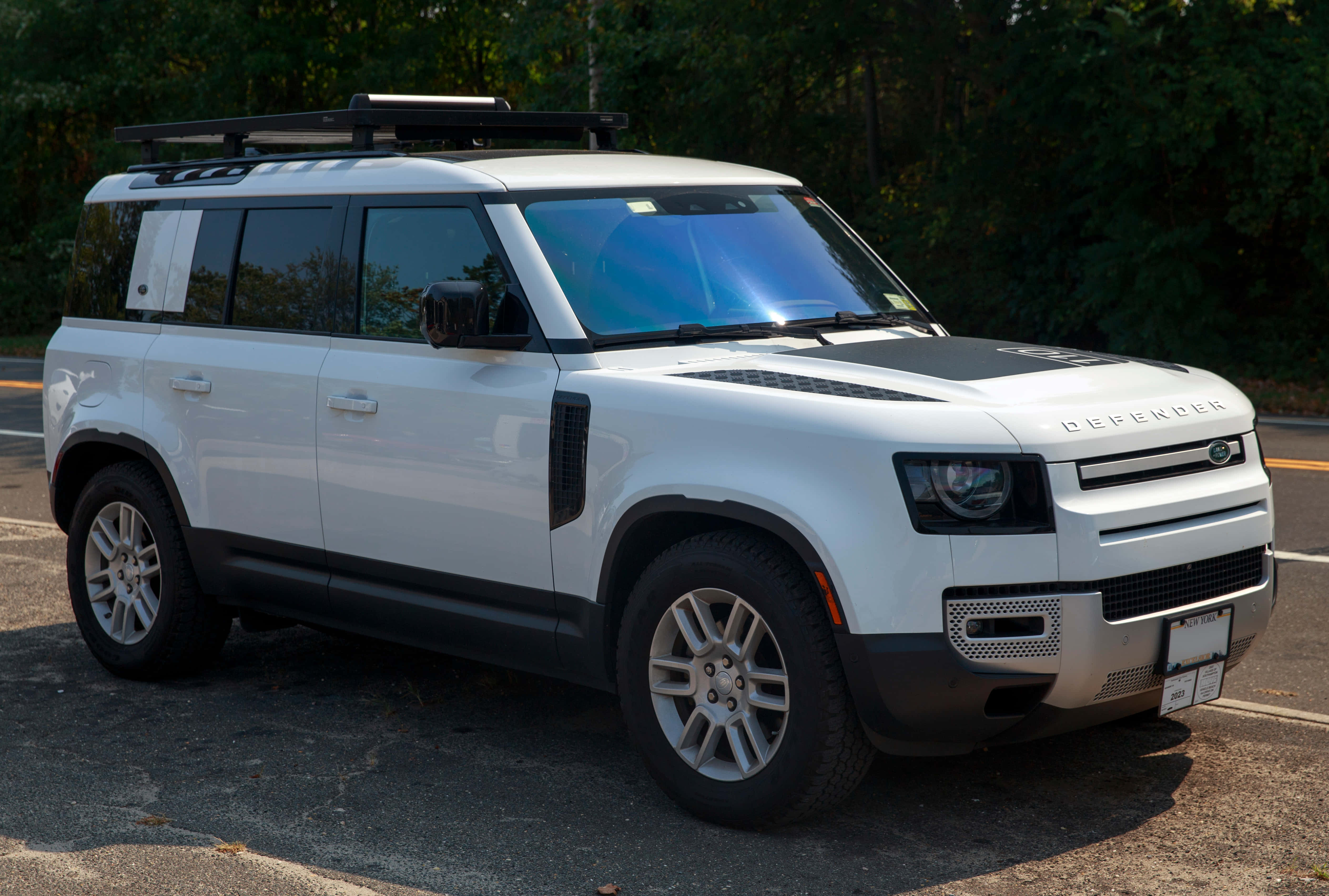 Land Rover Defender 桌布 壁紙 — 一輛純白色的 Land Rover Defender 停在瀝青路上，背景是茂密的綠樹，在明亮的日光下。
