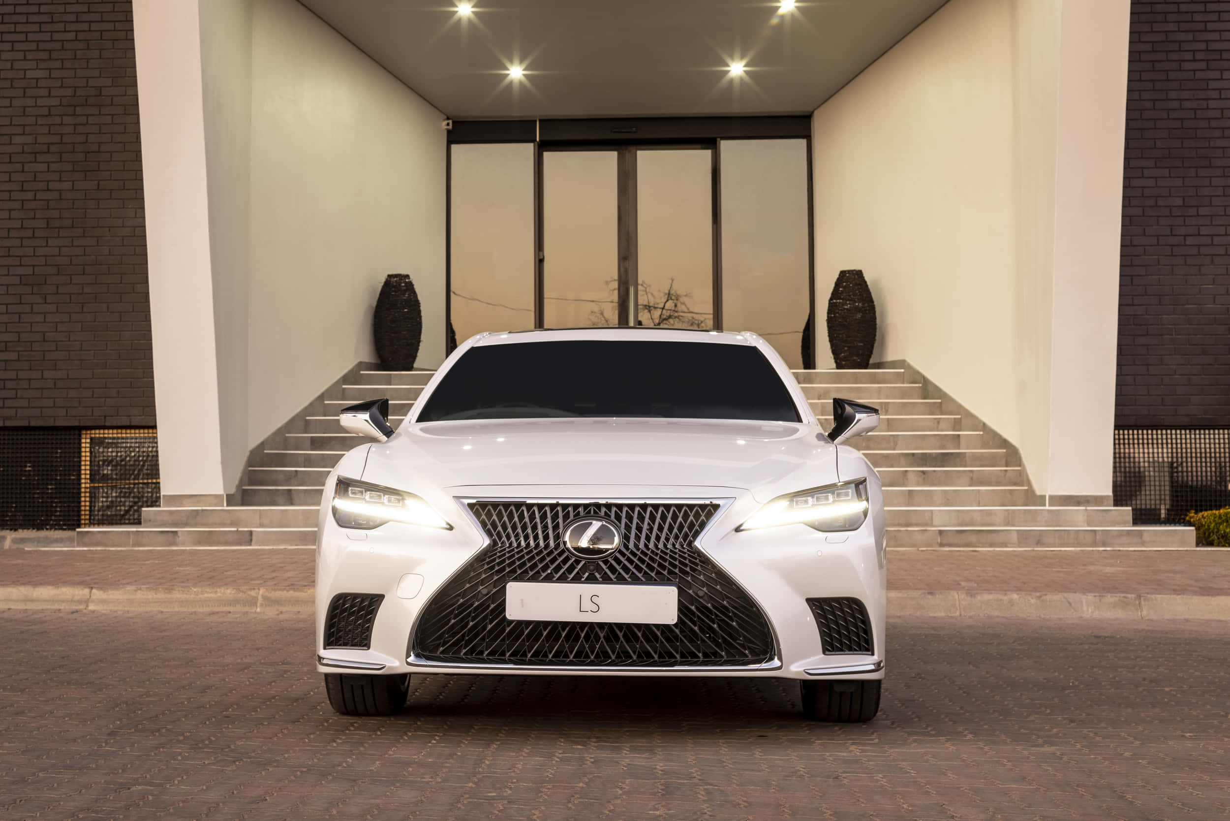 Lexus Ls 500 桌布 壁紙 — 一輛純白色的Lexus LS豪華轎車在明亮燈光下閃閃發光，停在帶有石階的現代建築入口前。