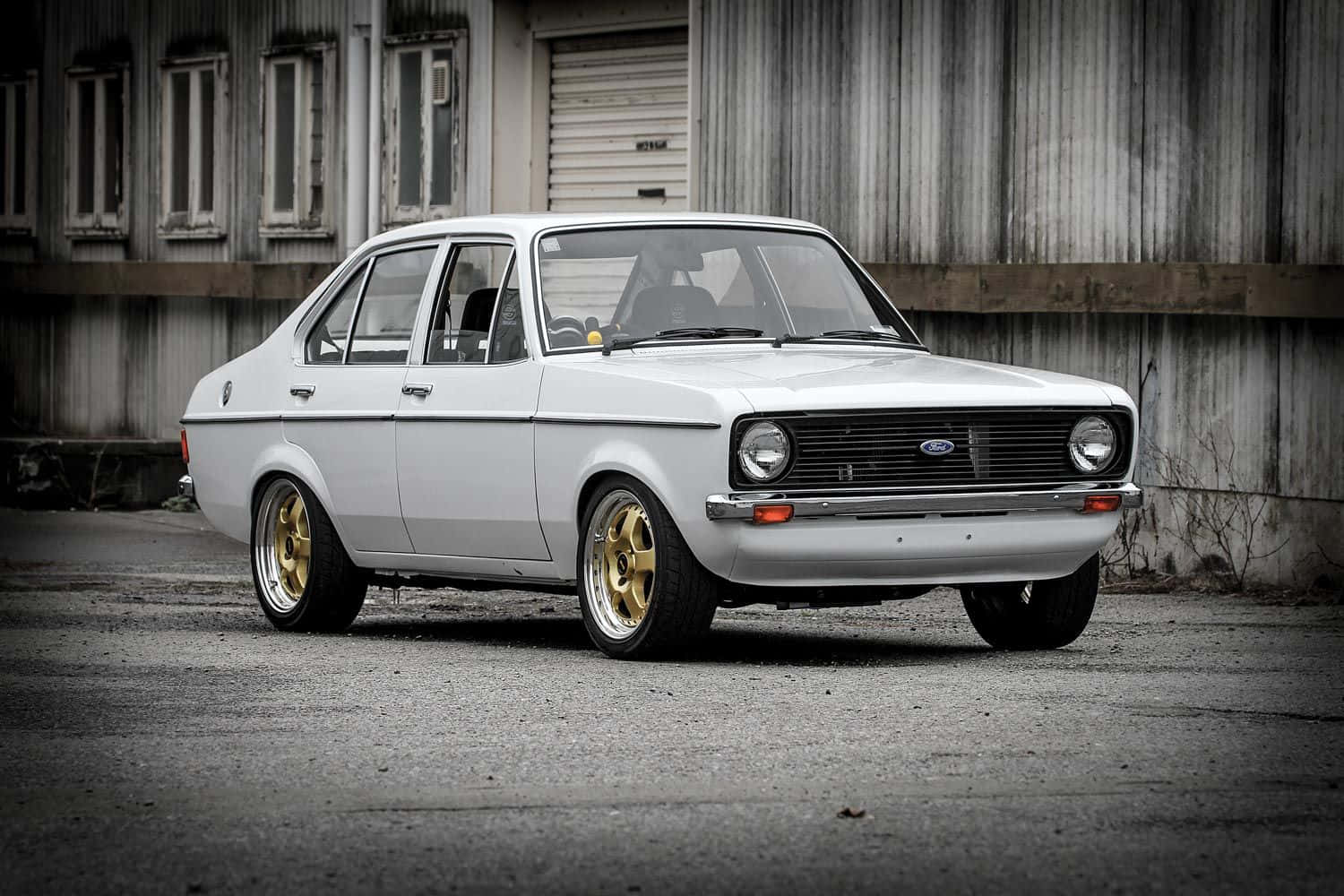 Ford Escort 桌布 壁紙 — 一輛純白色復古福特Escort，配金色輪圈，停泊在風化的工業建築前。