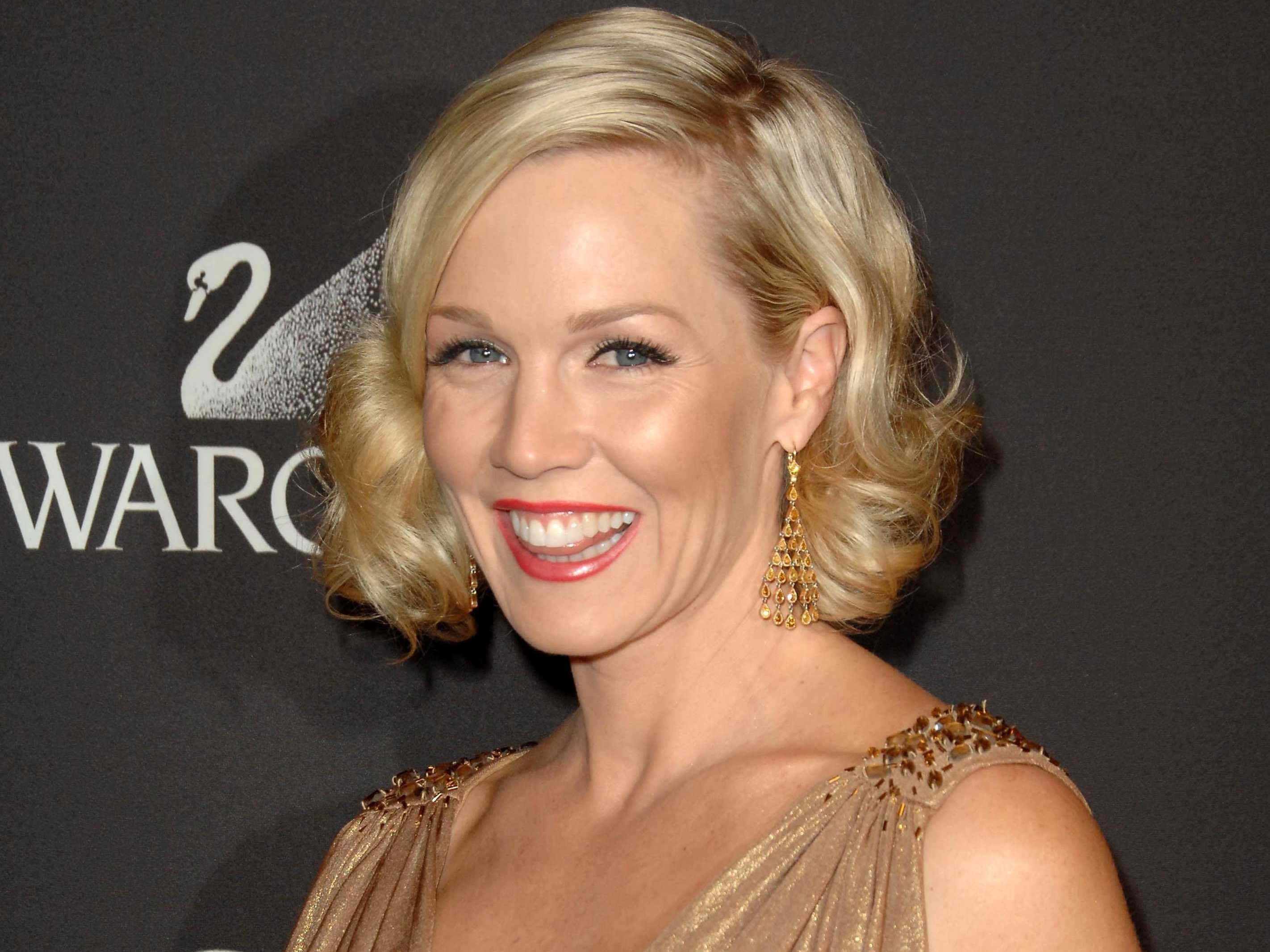 Jennie Garth 桌布 壁紙 — 一位光彩照人的金髮女性，塗著紅色唇膏，戴著金色耳環，穿著閃閃發光的金色連衣裙，在施華洛世奇頒獎典禮上微笑。