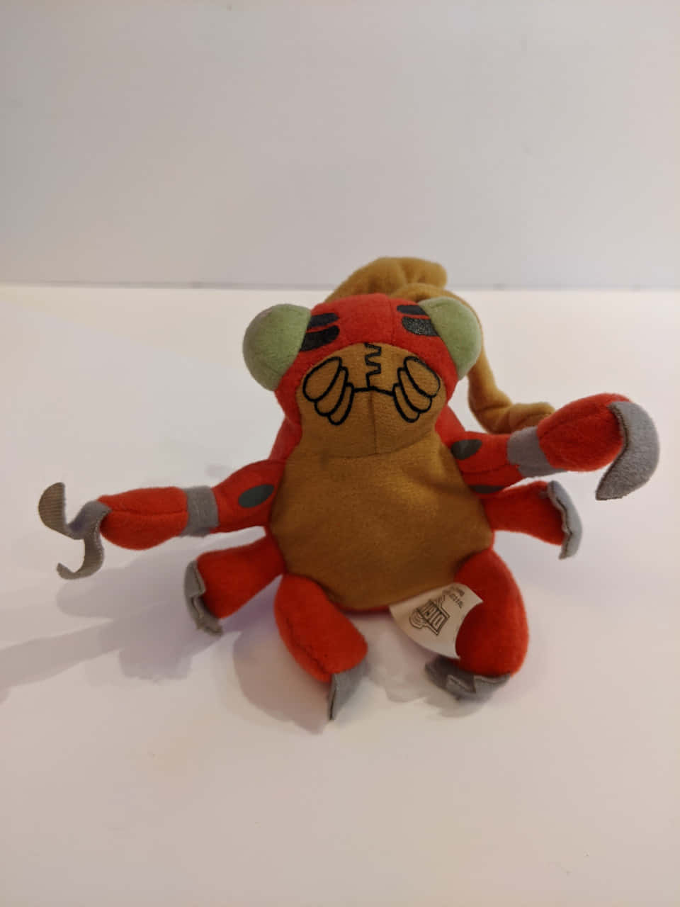 Digimon Tentomon 桌布 壁紙 — 一個紅色和橙色的毛絨玩具，有綠色耳朵和灰色爪子，坐在純白色的表面上。