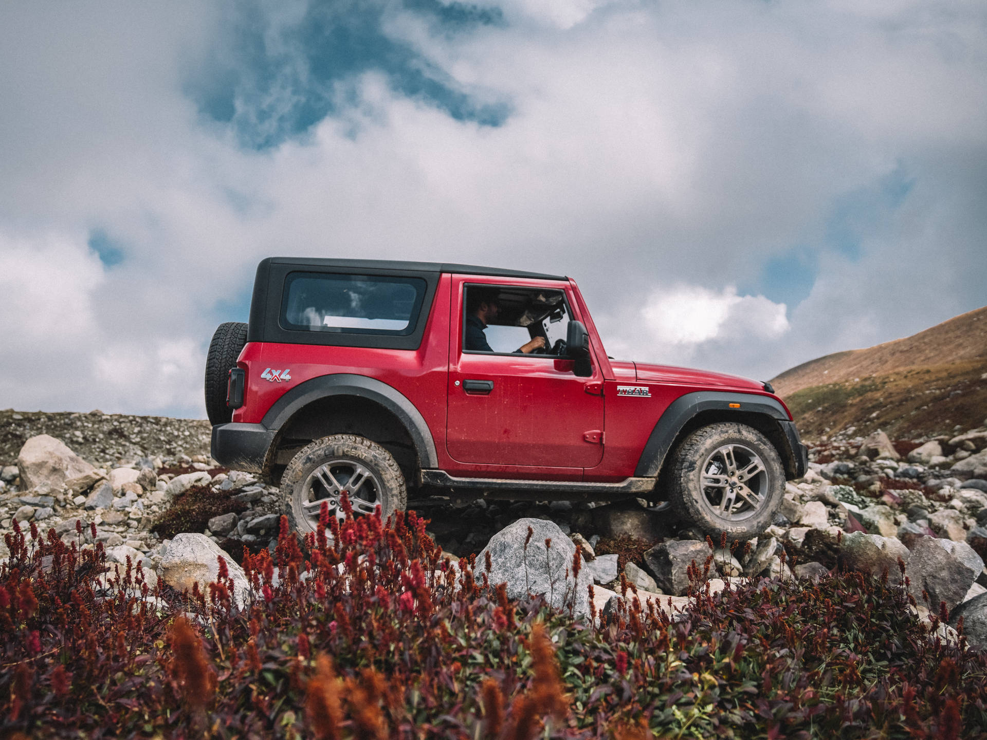 紅色Jeep Wrangler在岩石上行駛，有紅花和陰天。