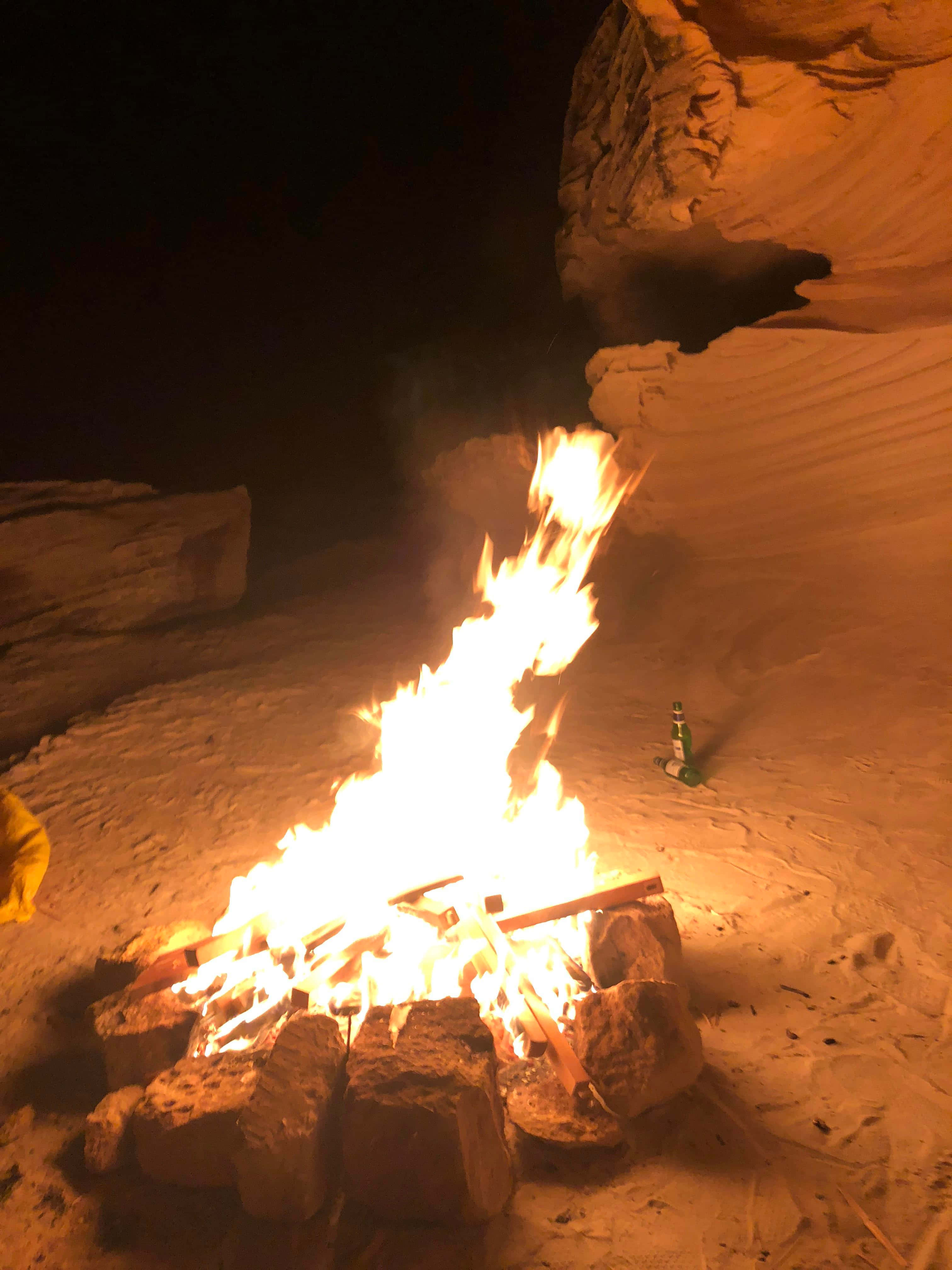 Beach Campfire 桌布 壁紙 — 一個咆哮的營火在沙漠峽谷中夜間明亮地燃燒，將溫暖的光線投射在砂岩牆壁和附近一個孤獨的瓶子上。