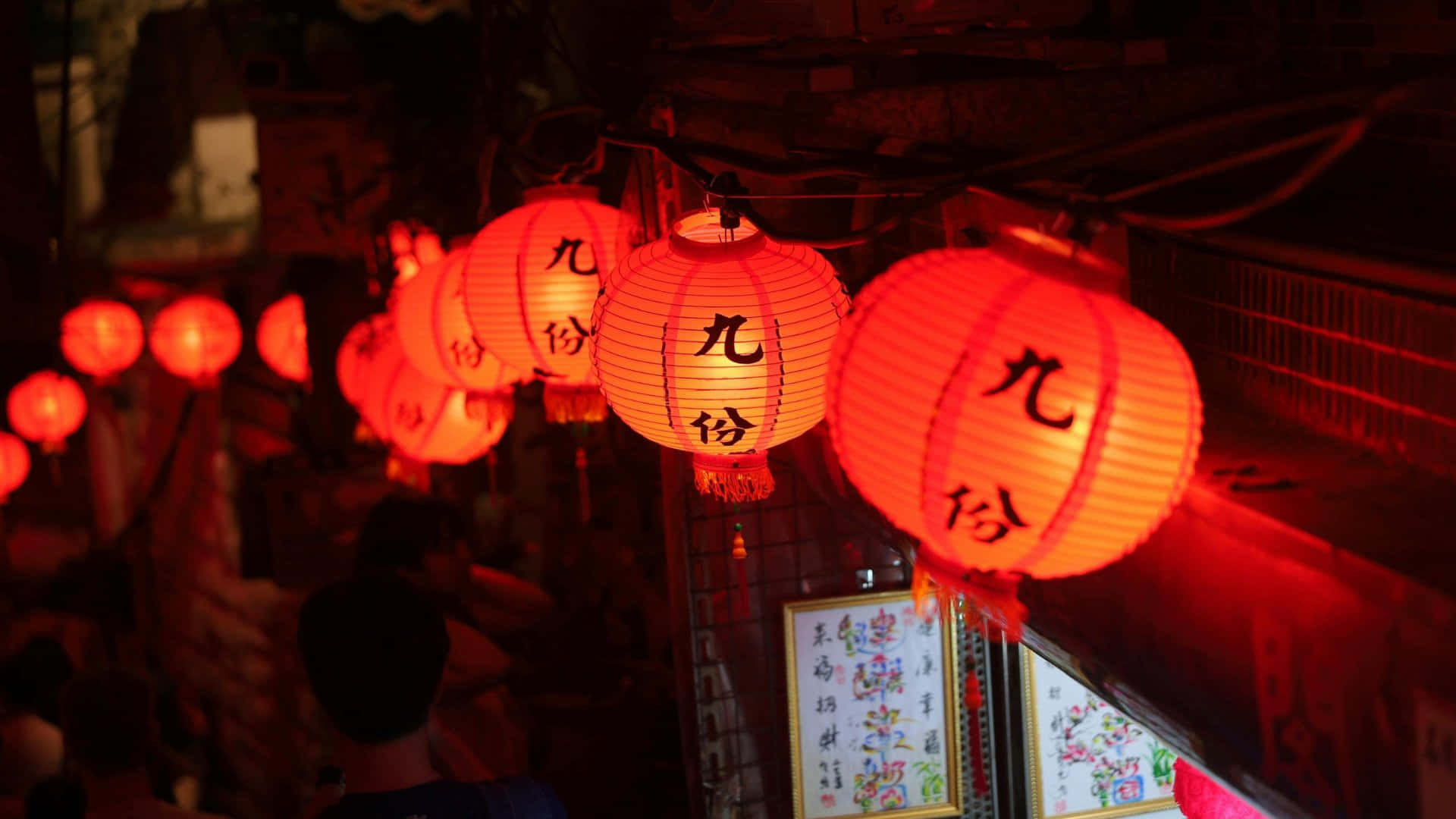  壁紙 — 一排發光的紅色紙燈籠，上面有黑色中文字，溫暖地懸掛在繁華的夜街上。