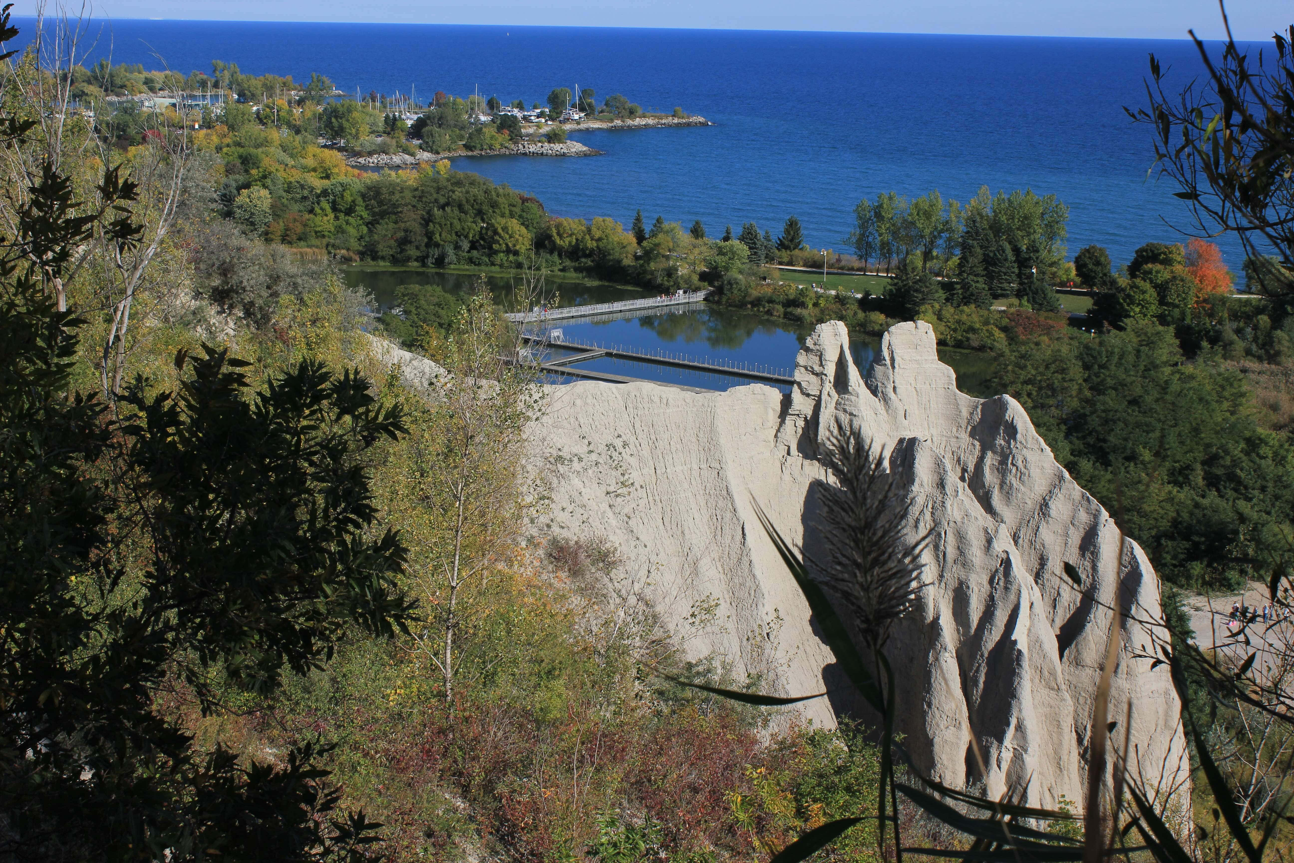Scarborough Bluffs 桌布 壁紙 — 一個風景優美的岩石懸崖俯瞰點，秋天的樹葉框著一座橋，橫跨平靜的海灣，通往晴朗天空下廣闊的藍色湖泊。