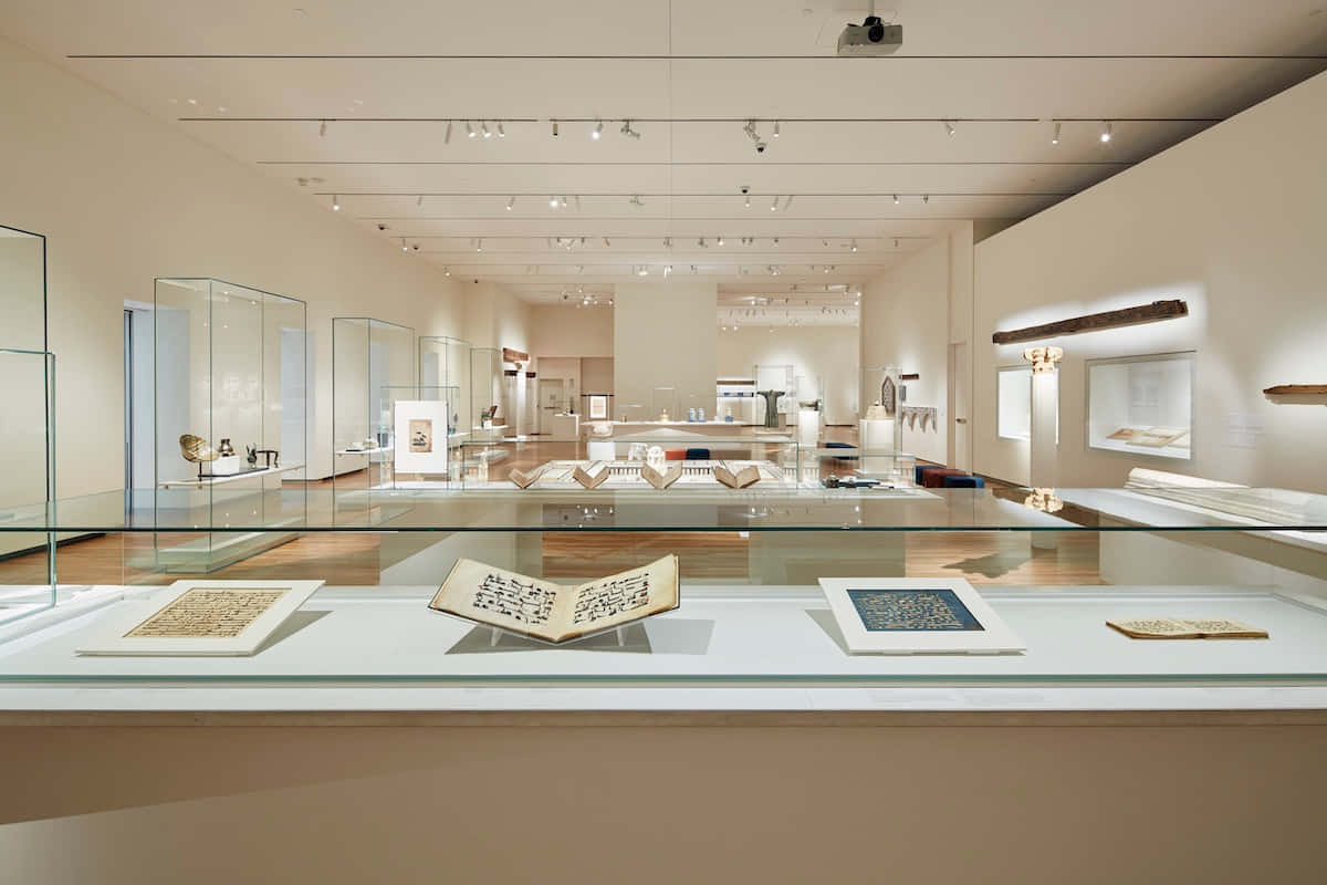 Aga Khan Museum 桌布 壁紙 — 一個寧靜的博物館畫廊，玻璃展示櫃在明亮的頭頂燈光下展示古代手稿和文物。