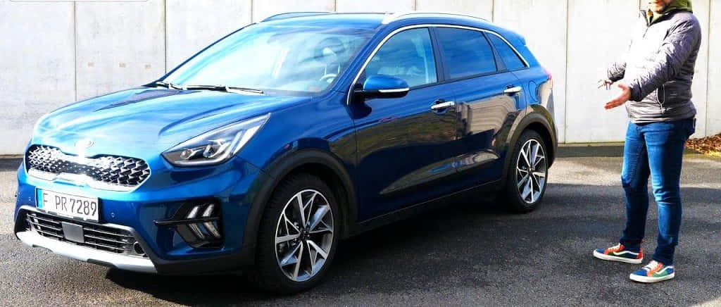 Kia Niro 桌布 壁紙 — 一架閃亮藍色Kia Niro SUV停泊在瀝青路上，旁邊有人做手勢，背景是樸素的混凝土牆。