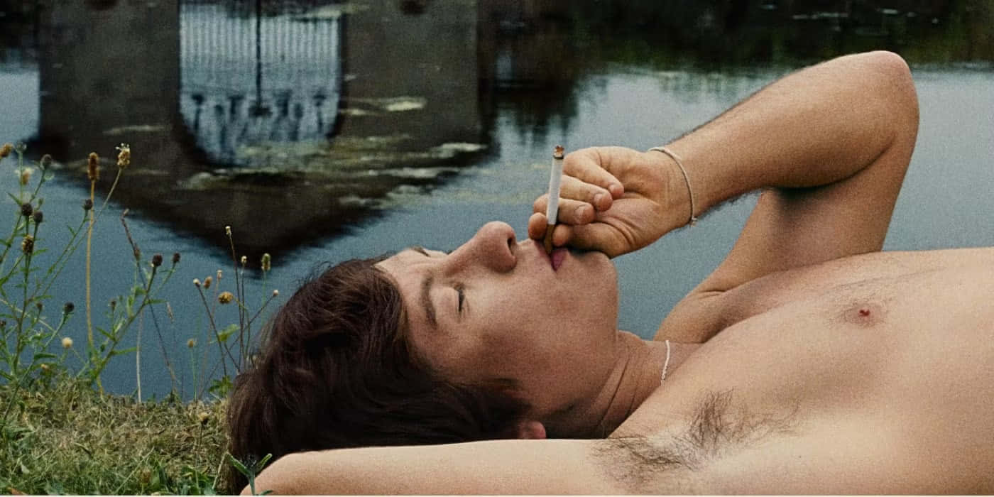  壁紙 — 一個赤裸上身的男人躺在水邊的草地上，閉著眼睛平靜地抽煙。