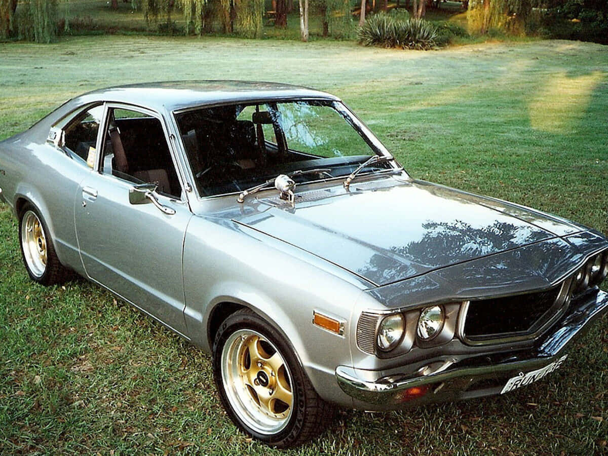 Mazda Rx-3 桌布 壁紙 — 一輛銀色經典雙門跑車配金色輪圈，停泊在陽光普照的草地上。