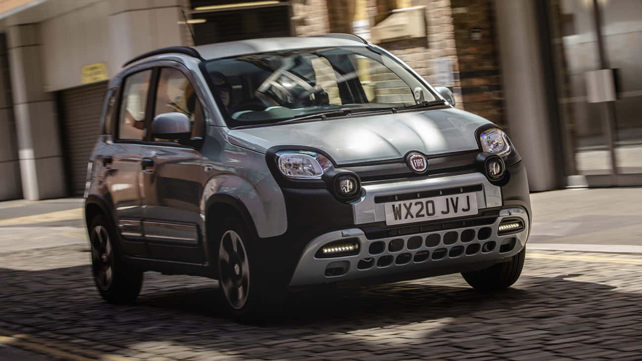 Fiat Panda 桌布 壁紙 — 一輛銀色嘅Fiat Panda喺鵝卵石街道上行駛，背景係模糊嘅城市建築。