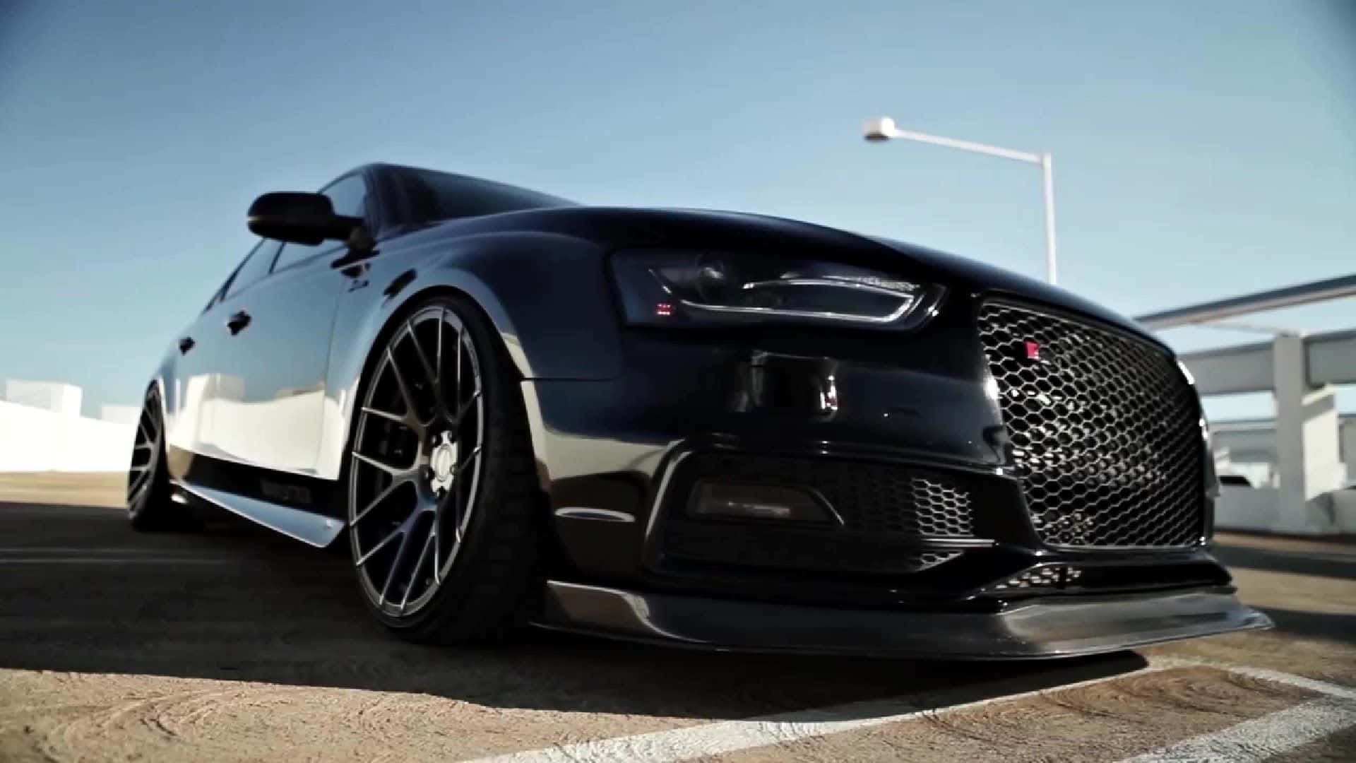 Audi S4 桌布 壁紙 — 一輛時尚的黑色奧迪轎車配備定制輪圈，停在陽光明媚的停車場，天空清澈湛藍。