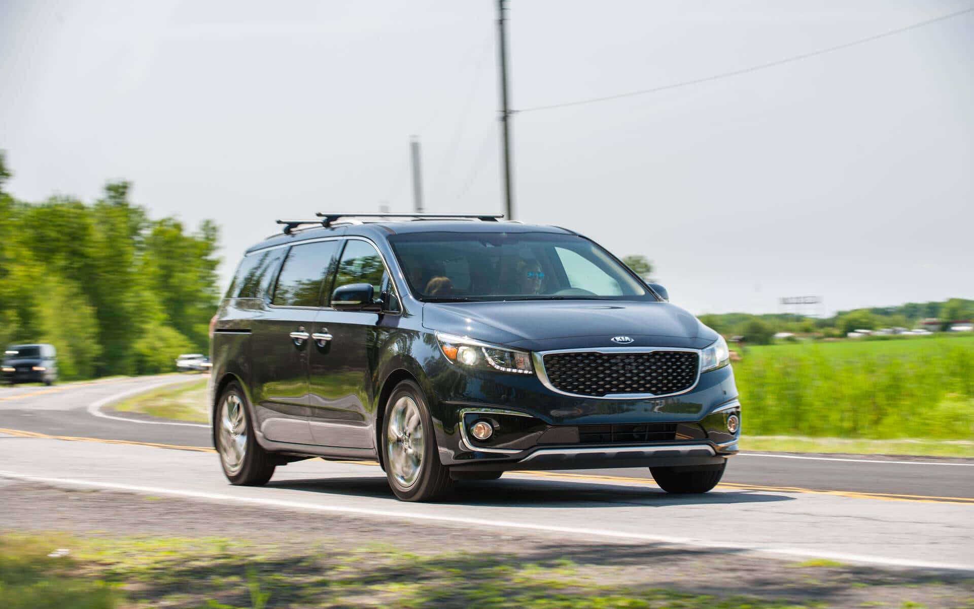 Kia Sedona 桌布 壁紙 — 一架時尚的黑色Kia小型貨車沿著陽光明媚的鄉村道路行駛，兩旁是茂密的綠樹和田野。