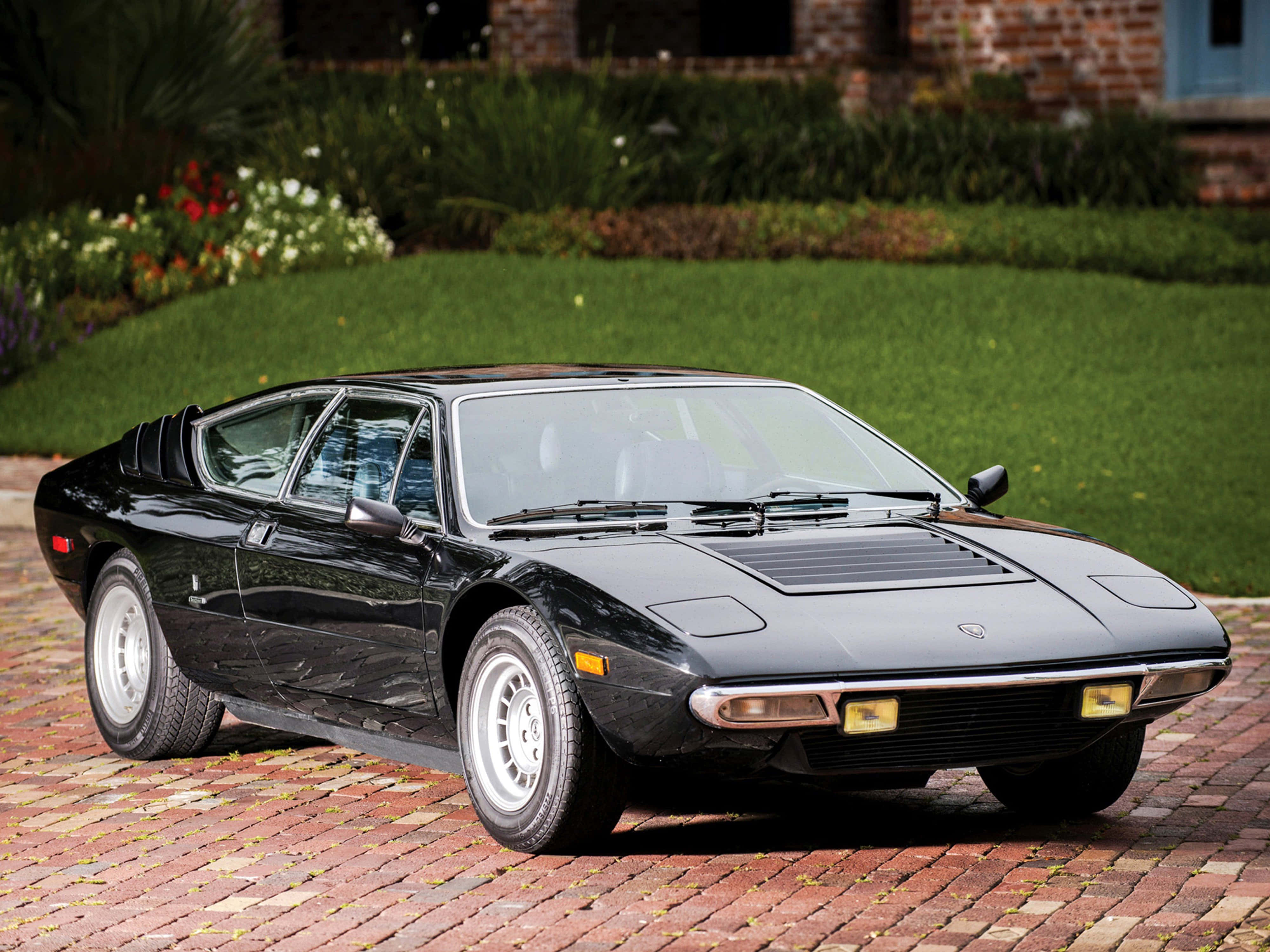 Lamborghini Urraco 桌布 壁紙 — 一架時尚的黑色林寶堅尼Countach停泊在紅磚車道上，背景是修剪整齊的草坪和磚屋。