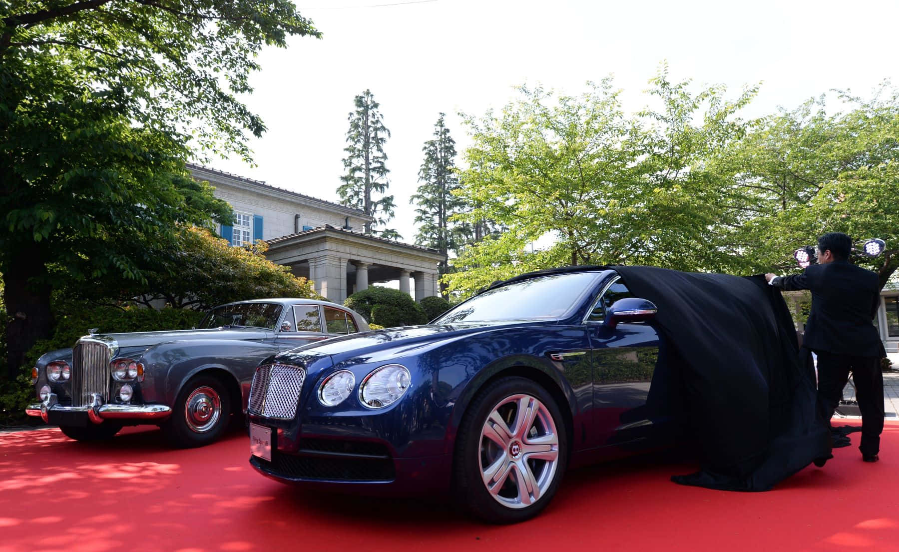 Bentley S3 桌布 壁紙 — 一架時尚的藍色賓利在紅地毯上由一名穿黑衣的男子揭幕，旁邊有一輛復古車在明亮的日光下。