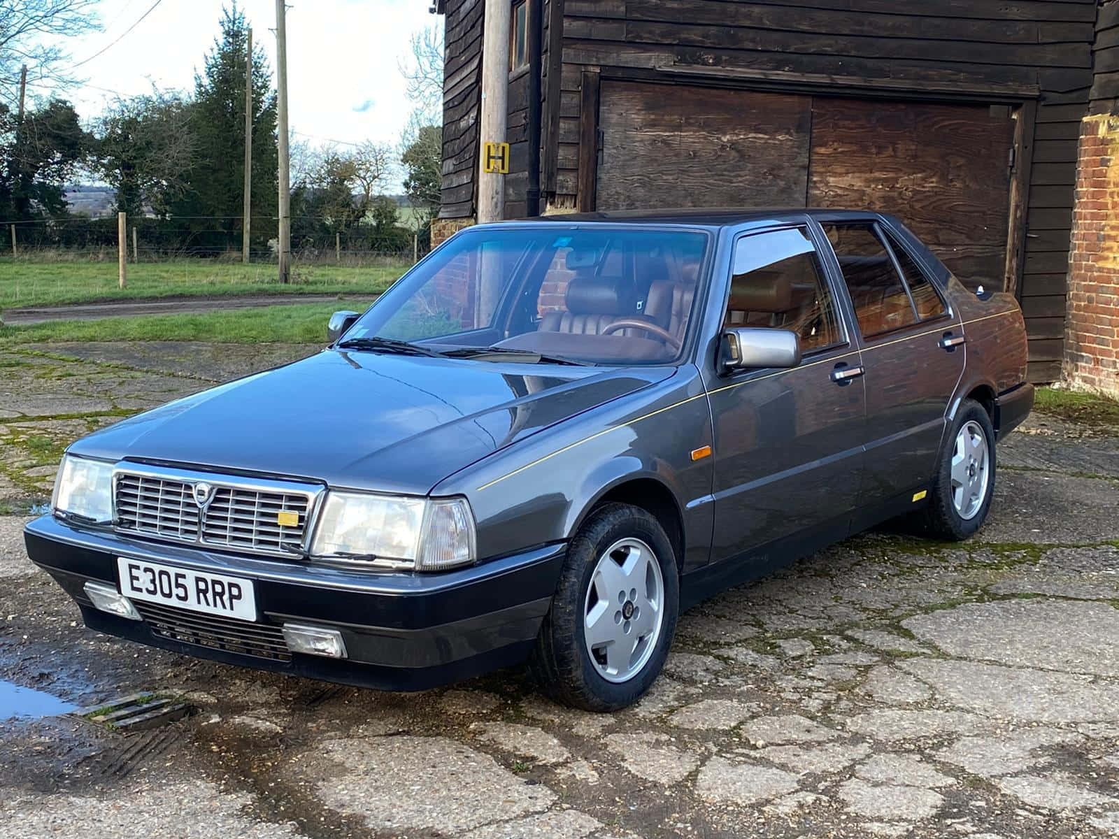  壁紙 — 一架時尚深灰色 Lancia Thema 房車停泊在裂開的石頭車道上，旁邊是質樸的木車庫。