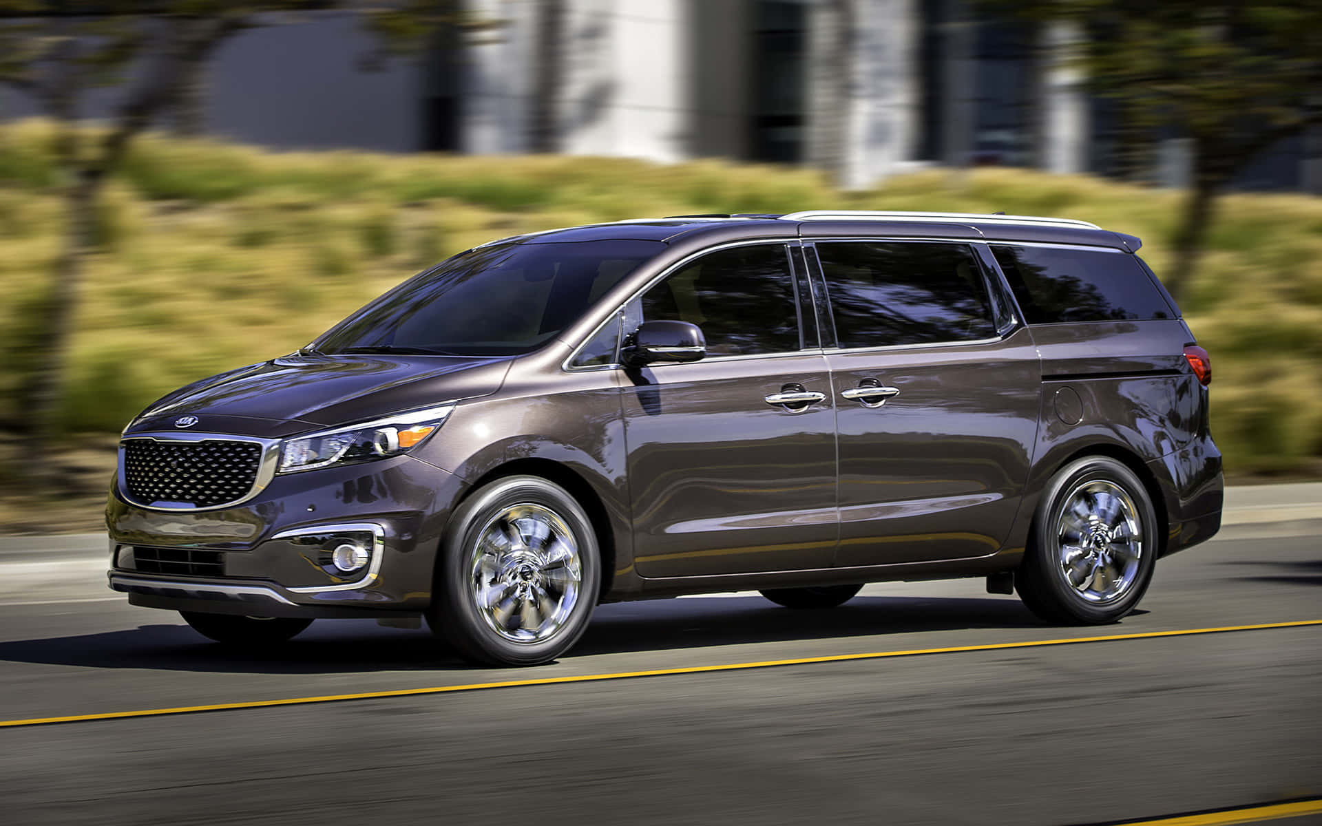Kia Sedona 桌布 壁紙 — 一架時尚的深灰色小型貨車在陽光明媚的道路上行駛，背景有模糊的綠化和建築物。