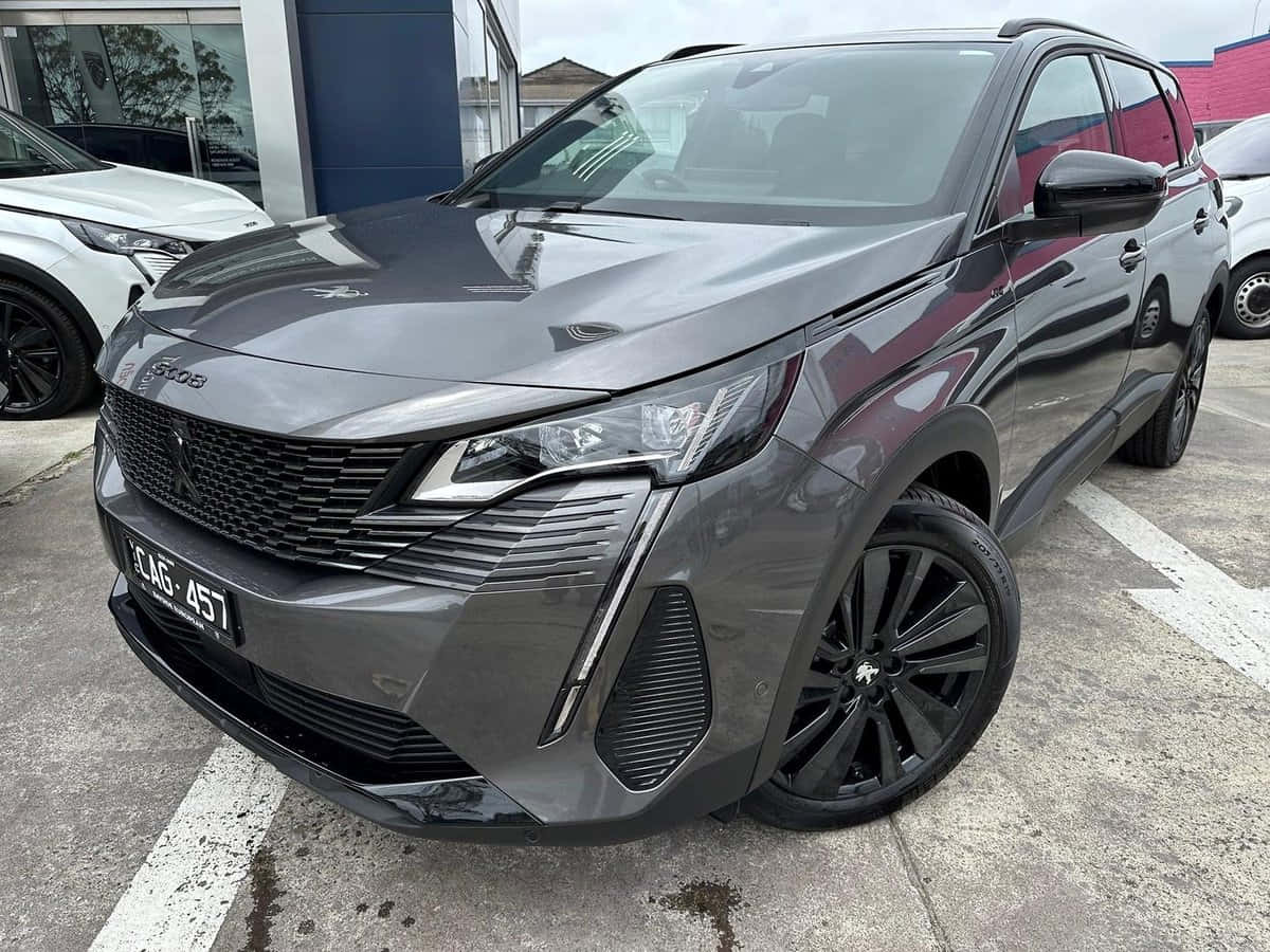 Peugeot 5008 桌布 壁紙 — 一輛時尚的深灰色標緻SUV停放在經銷商停車場，附近有現代建築和其他車輛。