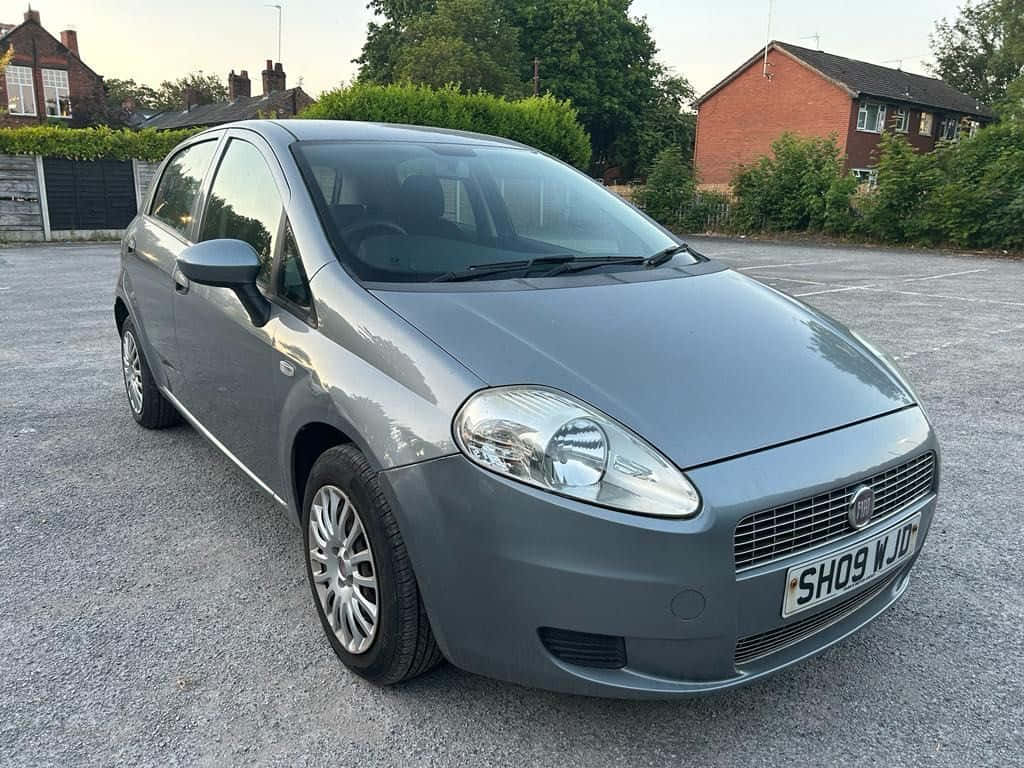 Fiat Punto 桌布 壁紙 — 一輛時尚的灰色 Fiat Punto 停泊在碎石地上，背景是黃昏時分的磚房和綠色籬笆。
