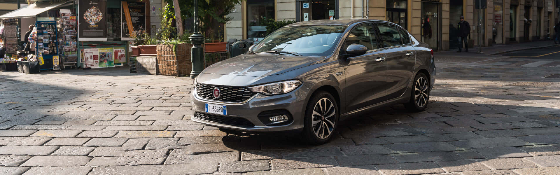 Fiat Tipo 桌布 壁紙 — 一架流線型灰色快意房車停泊在歐洲小鎮陽光普照的鵝卵石街道上，附近有商店和行人。