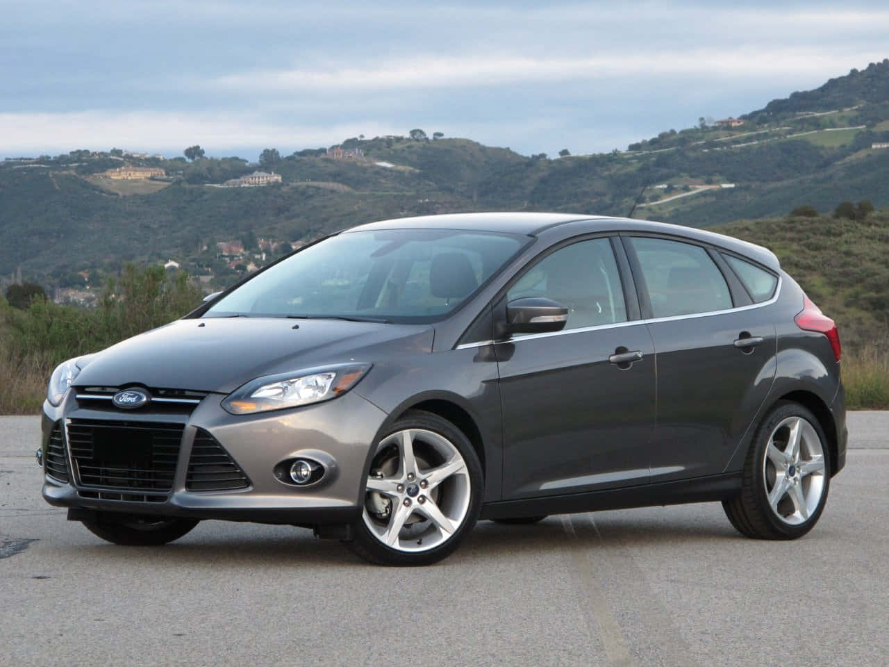Ford Focus 桌布 壁紙 — 一架時尚的灰色福特福克斯掀背車停在鋪砌的道路上，背景是起伏的綠色山丘和散落的房屋，天空多雲。