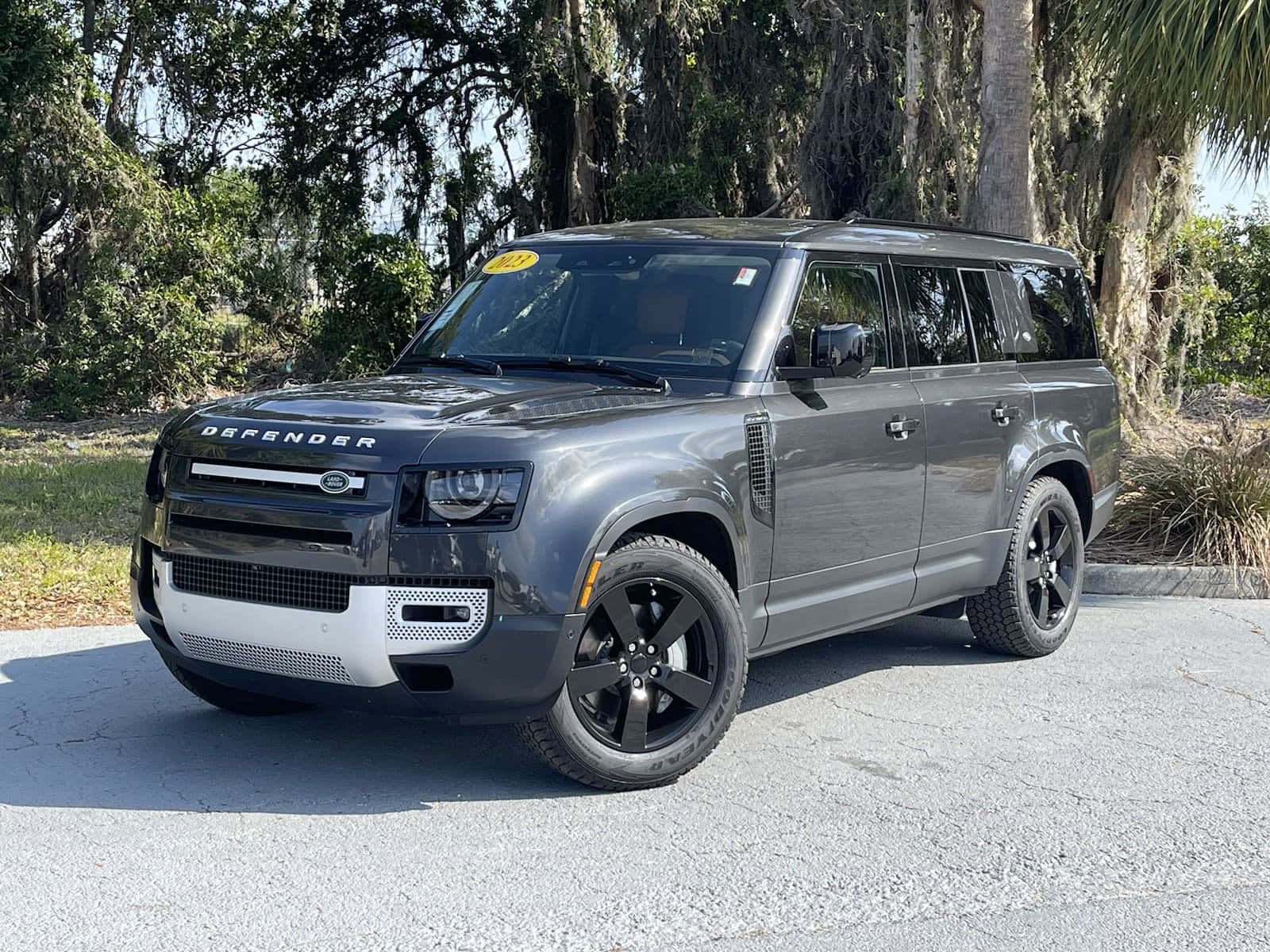 Land Rover Defender 桌布 壁紙 — 一輛時尚的灰色 Land Rover Defender SUV 停泊在裂開的路面上，背景是明亮的日光和茂密的綠樹。