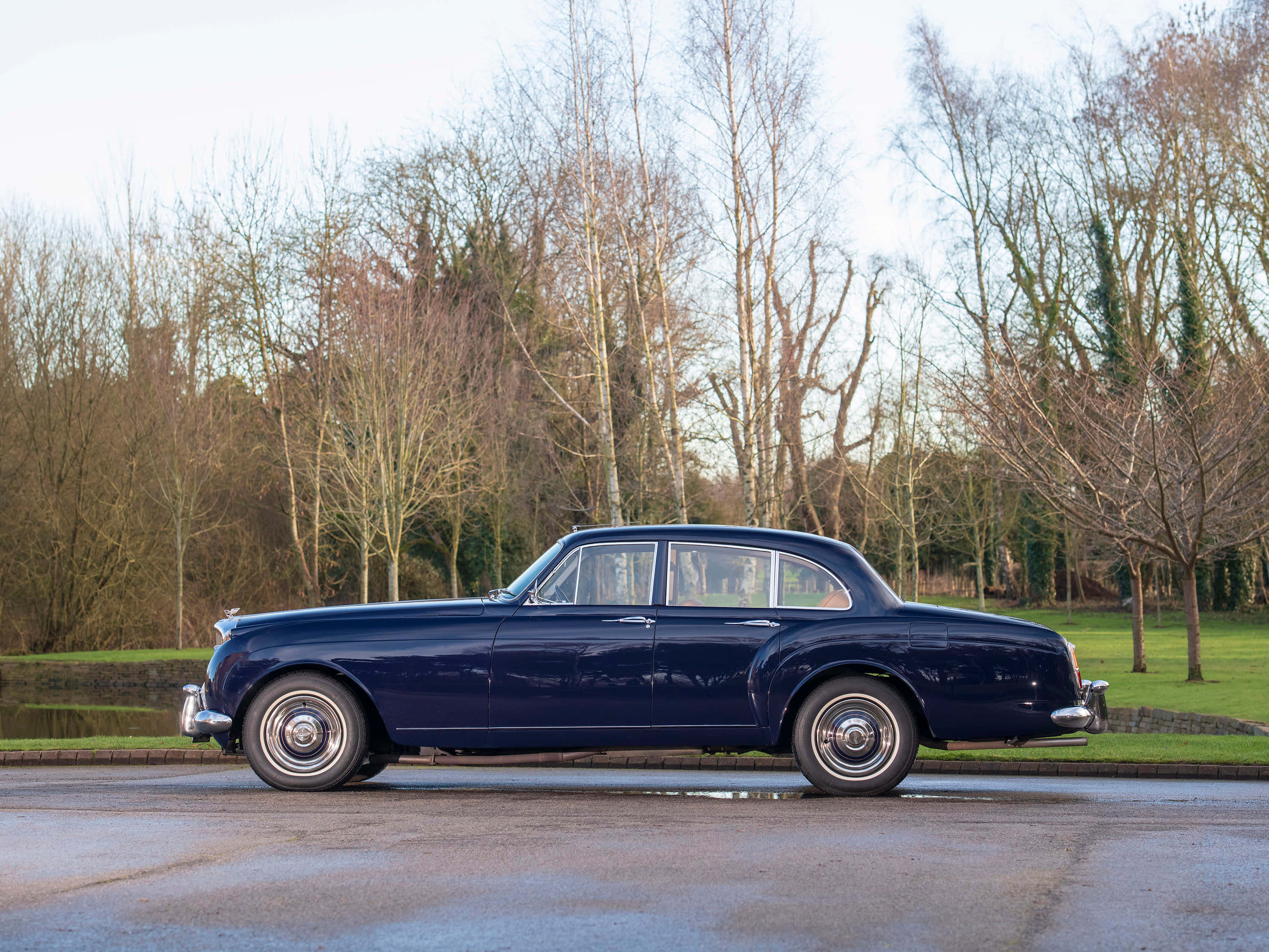 Bentley S2 桌布 壁紙 — 一輛時尚的海軍藍經典汽車在柔和日光下閃耀，停在濕路上，旁邊是無葉的樹木和草坡。