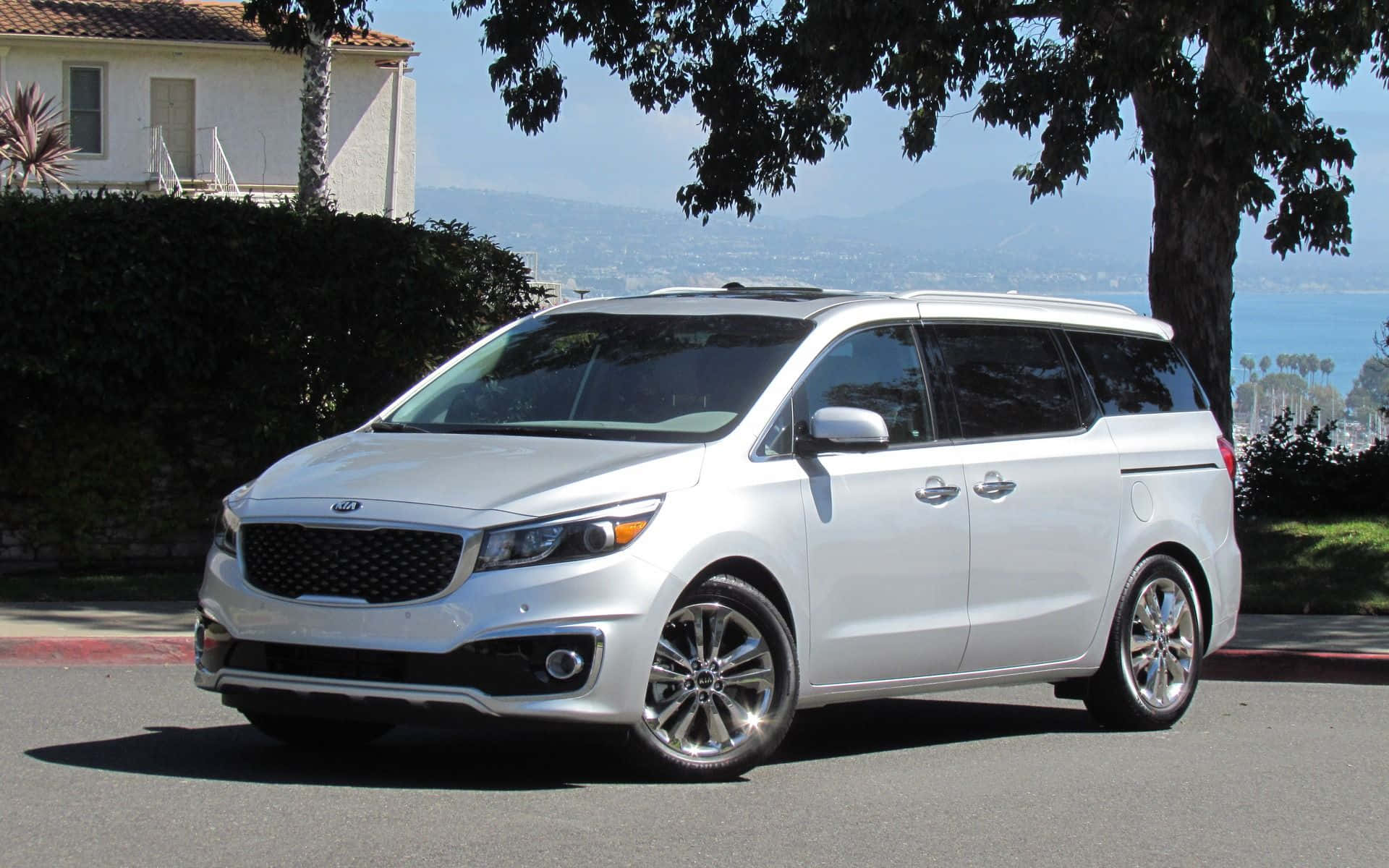 Kia Sedona 桌布 壁紙 — 一架時尚的銀色Kia小型貨車停泊在陽光明媚的街道上，背景有樹木、遠山和海洋。