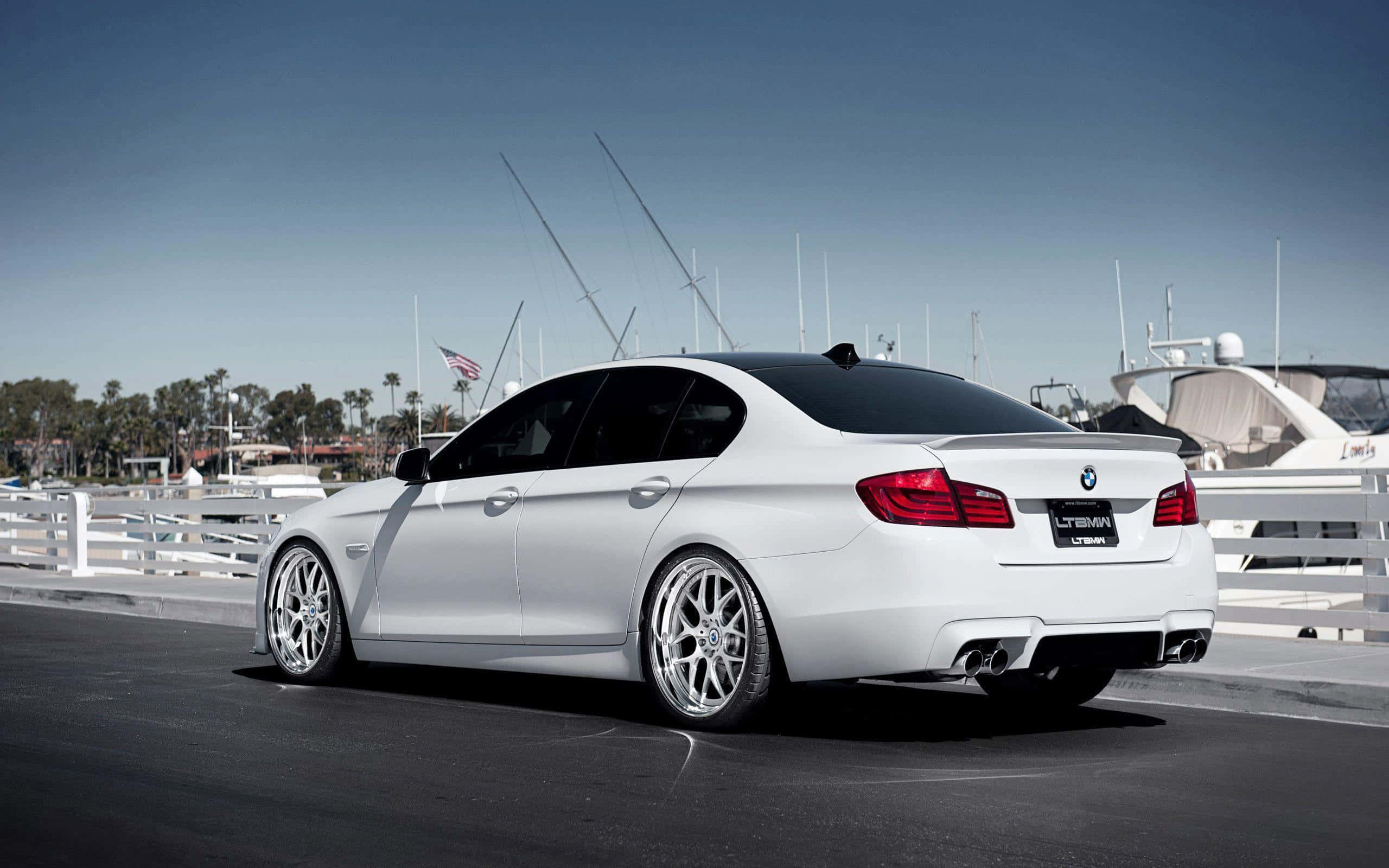 Bmw M5 桌布 壁紙 — 一架時尚的白色BMW房車配備定制輪圈，停泊在碼頭旁，旁邊是豪華遊艇，天空清澈湛藍。