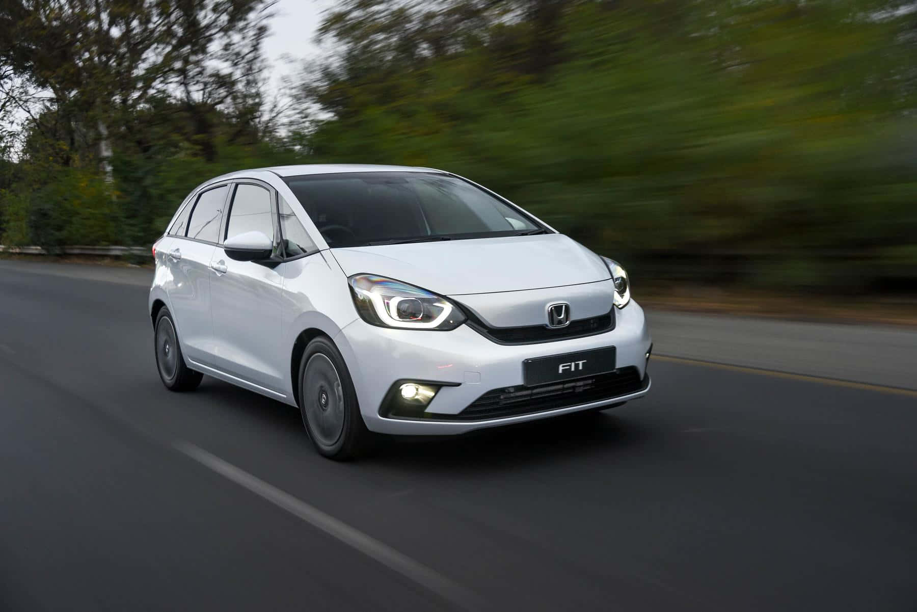 Honda Fit 桌布 壁紙 — 一輛時尚的白色本田飛度在綠樹成蔭的道路上行駛，動態模糊強調了其動態運動。