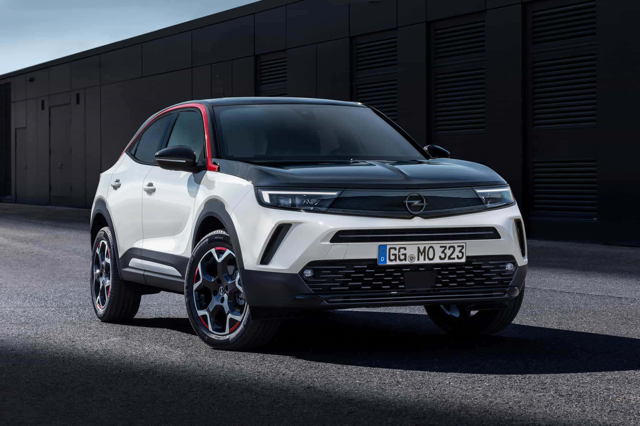 Opel Mokka 桌布 壁紙 — 一輛時尚的白色歐寶SUV，帶有紅色點綴，停在黑暗的工業建築前，在明亮的日光下。