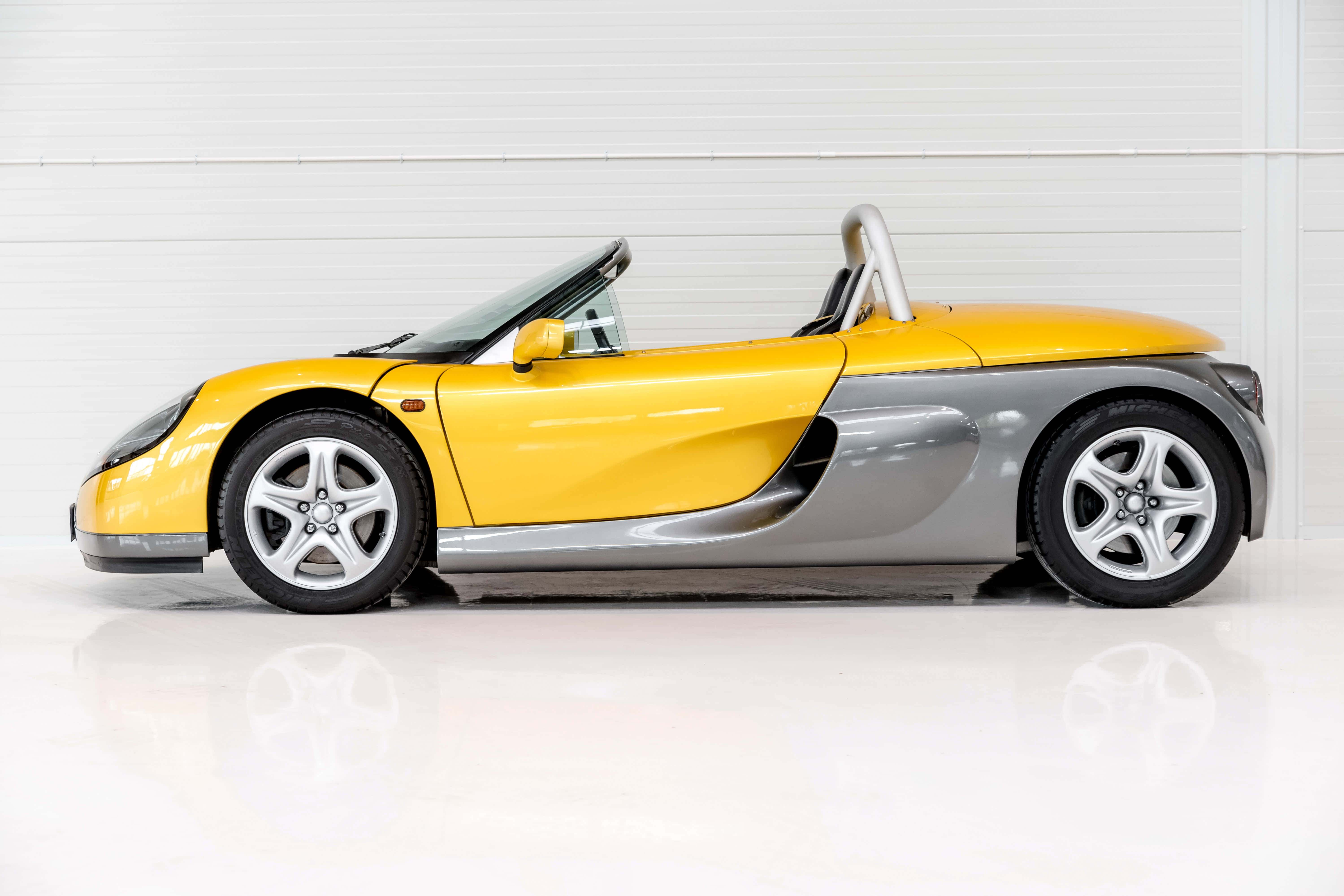 Renault Sport Spider 桌布 壁紙 — 一輛時尚的黃銀色敞篷跑車在明亮白色、帶反射地板的攝影棚中展示。