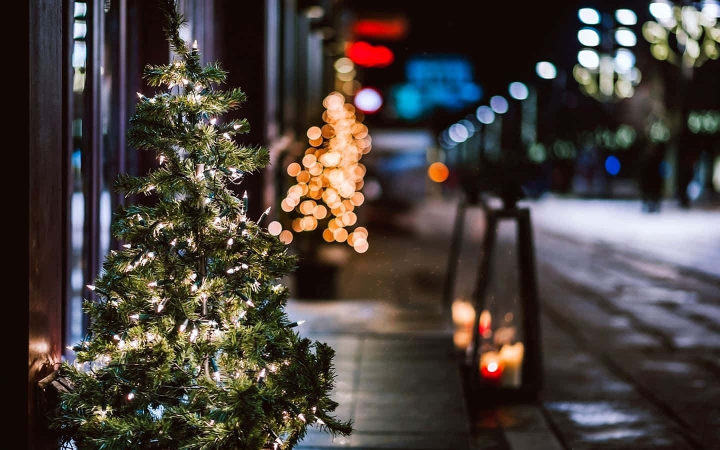  壁紙 — 一棵細小裝飾的聖誕樹在雪夜的城市行人路上閃爍著燈串，背景是散景的路燈。