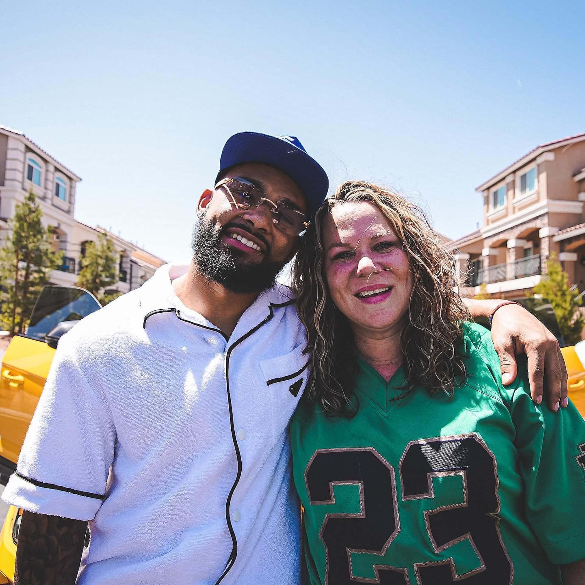  壁紙 — 一位微笑的男士和女士在室外一輛黃色汽車前合影，身後是住宅建築，頭頂是晴朗的藍天。