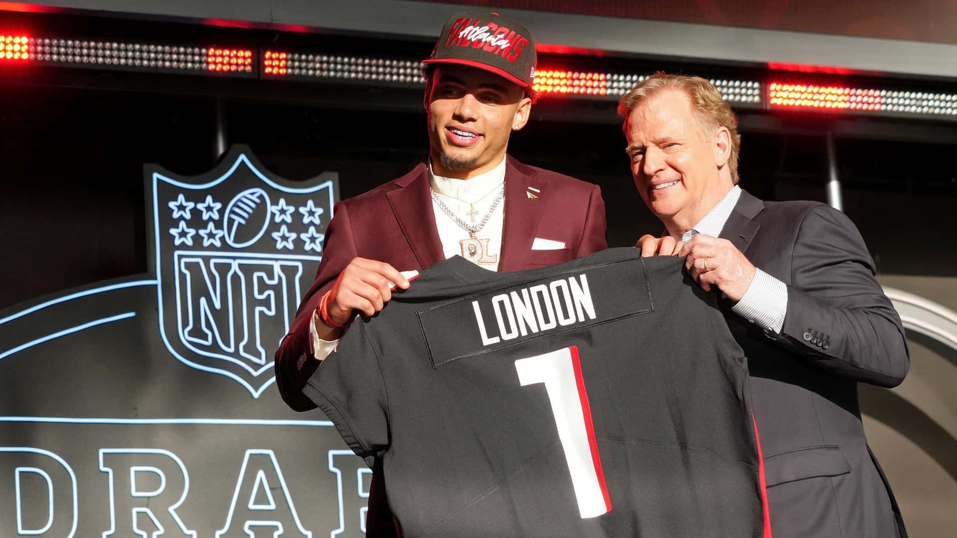  壁紙 — 一位微笑的NFL選秀球員身穿栗色西裝，舉起印有'LONDON'的黑色一號球衣，站在微笑的官員旁邊，背景是閃亮的NFL選秀舞台。