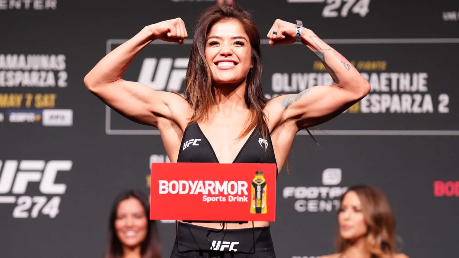  壁紙 — 一位微笑的UFC拳手在帶有'UFC 274'品牌和BodyArmor標誌的稱重台上彎曲她肌肉發達的手臂。