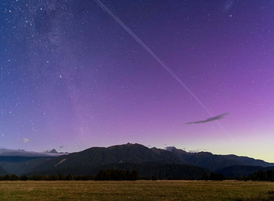  壁紙 — 一個繁星點點的紫色暮色天空，帶有飛機軌跡，拱形在剪影山脈和草地之上。