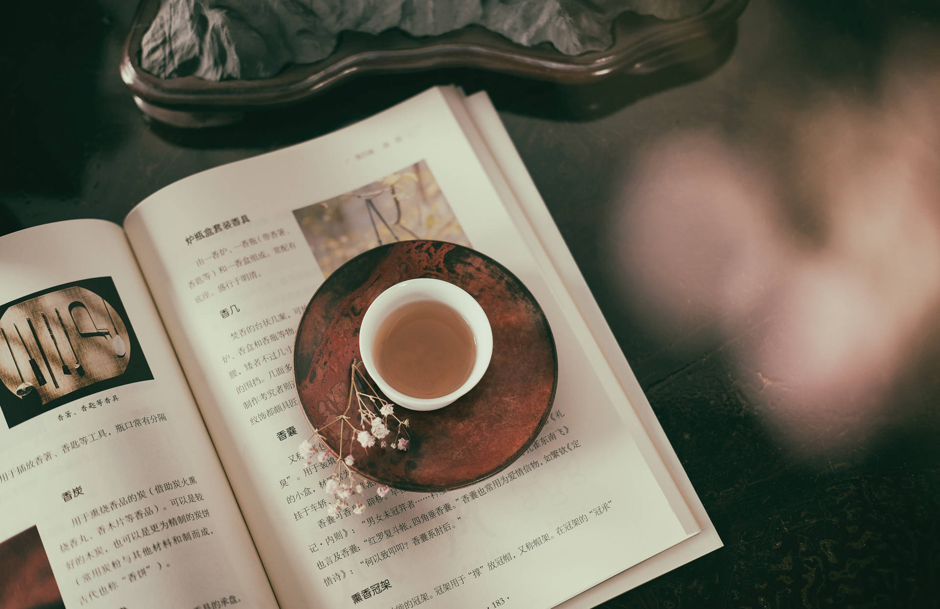 中式平鋪攝影 壁紙 — 一杯冒煙的茶放在木製茶托上，擱在一本打開的書上，周圍是柔和自然光線和模糊的樹葉。