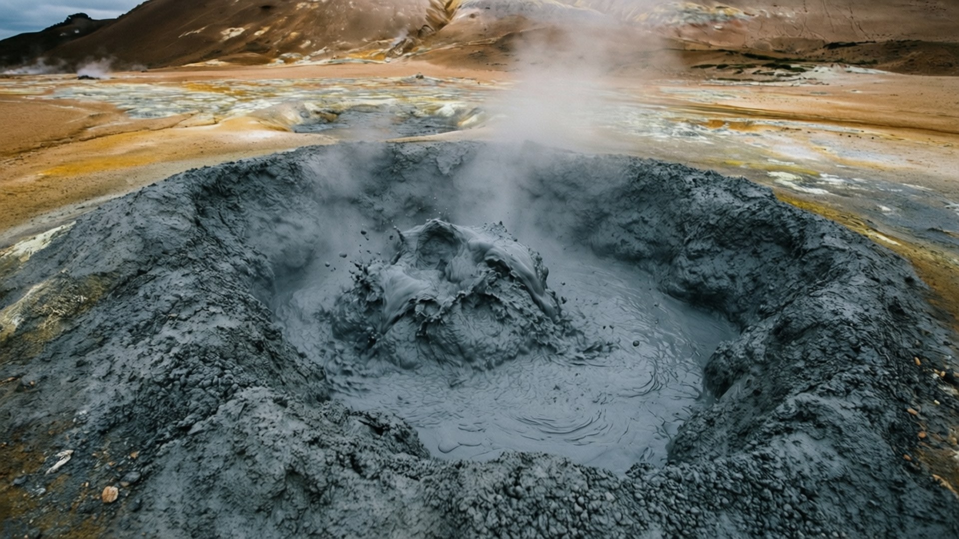 Rotorua 桌布 壁紙 — 一個冒著蒸汽的火山泥鍋在陰天之下，於崎嶇、大地色調的地熱景觀中冒泡翻騰。