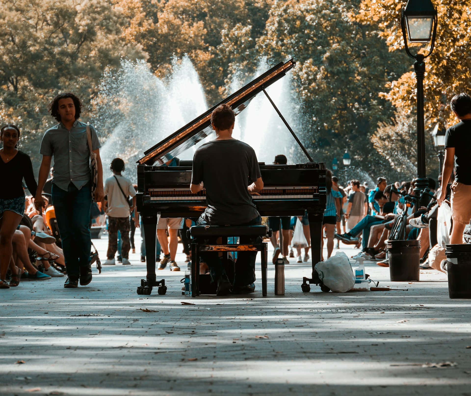  壁紙 — 一位街頭音樂家在陽光普照的公園裡彈奏三角鋼琴，人們在噴泉周圍漫步和坐著。