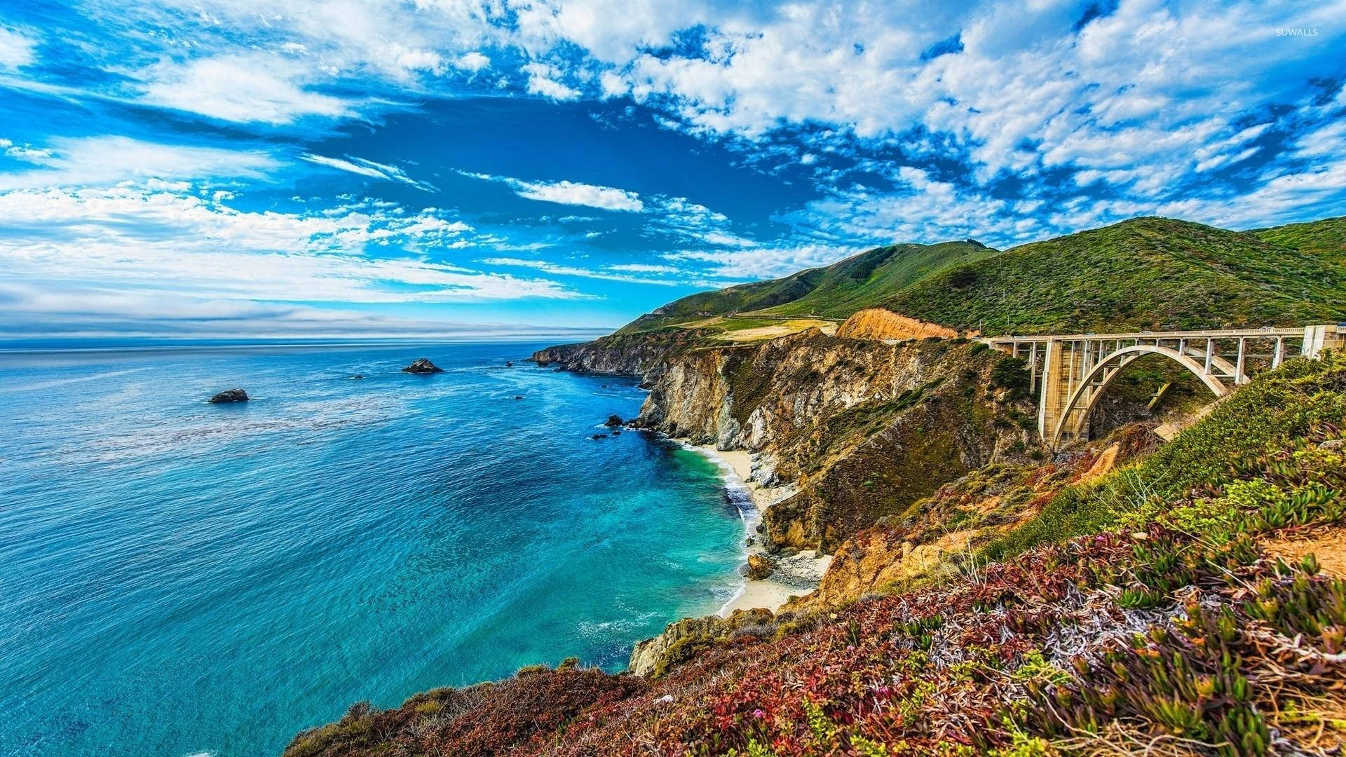 Big Sur陰天海灘 壁紙 — 壯麗的海岸景觀，標誌性的比克斯比橋橫跨崎嶇的懸崖，下方是碧綠的海水，上方是湛藍的天空和蓬鬆的白雲。