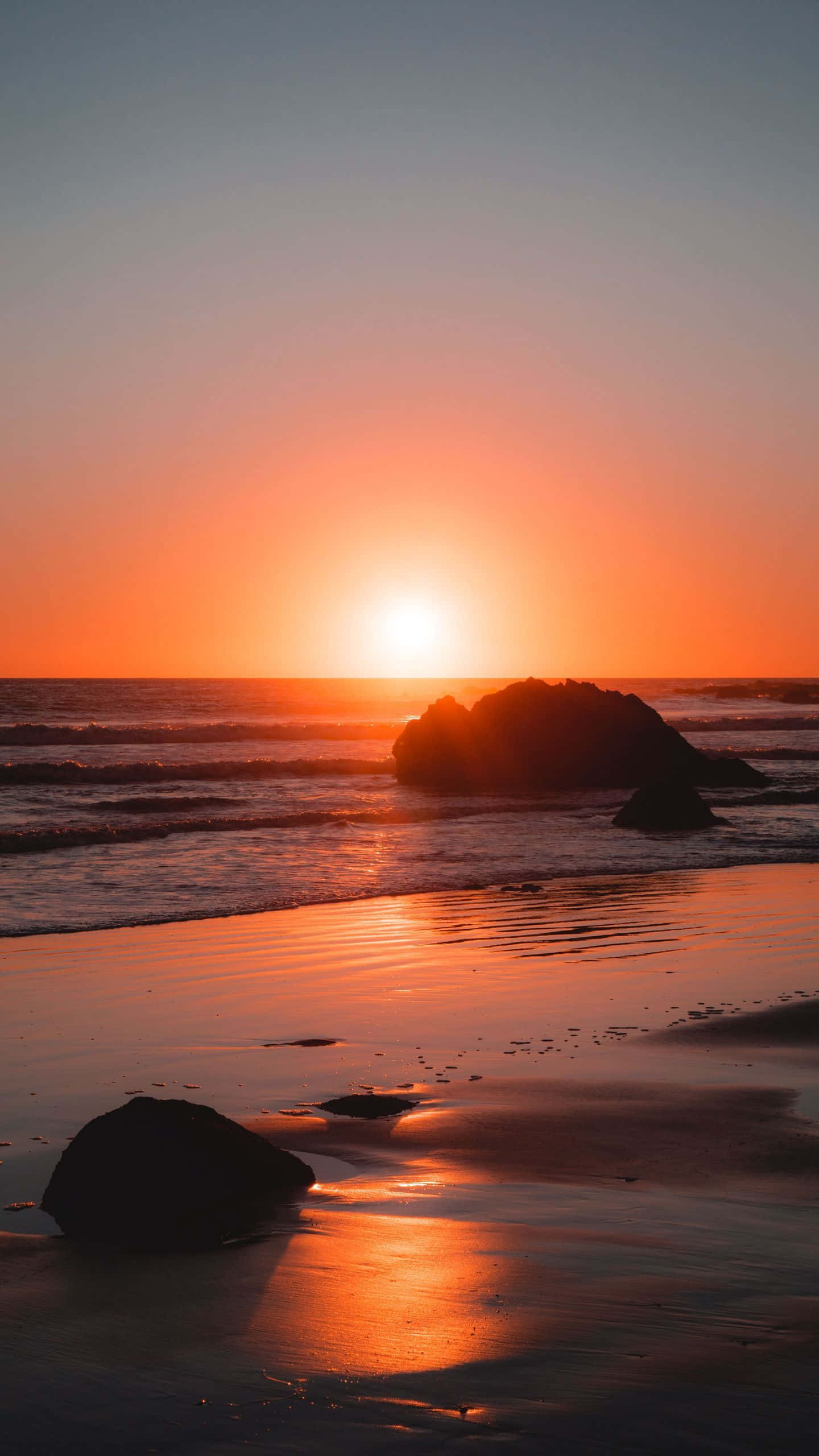 Coastal Sunset 桌布 壁紙 — 寧靜海洋上壯麗的日落，發光的橙色光線反射在濕沙和剪影岩石上。