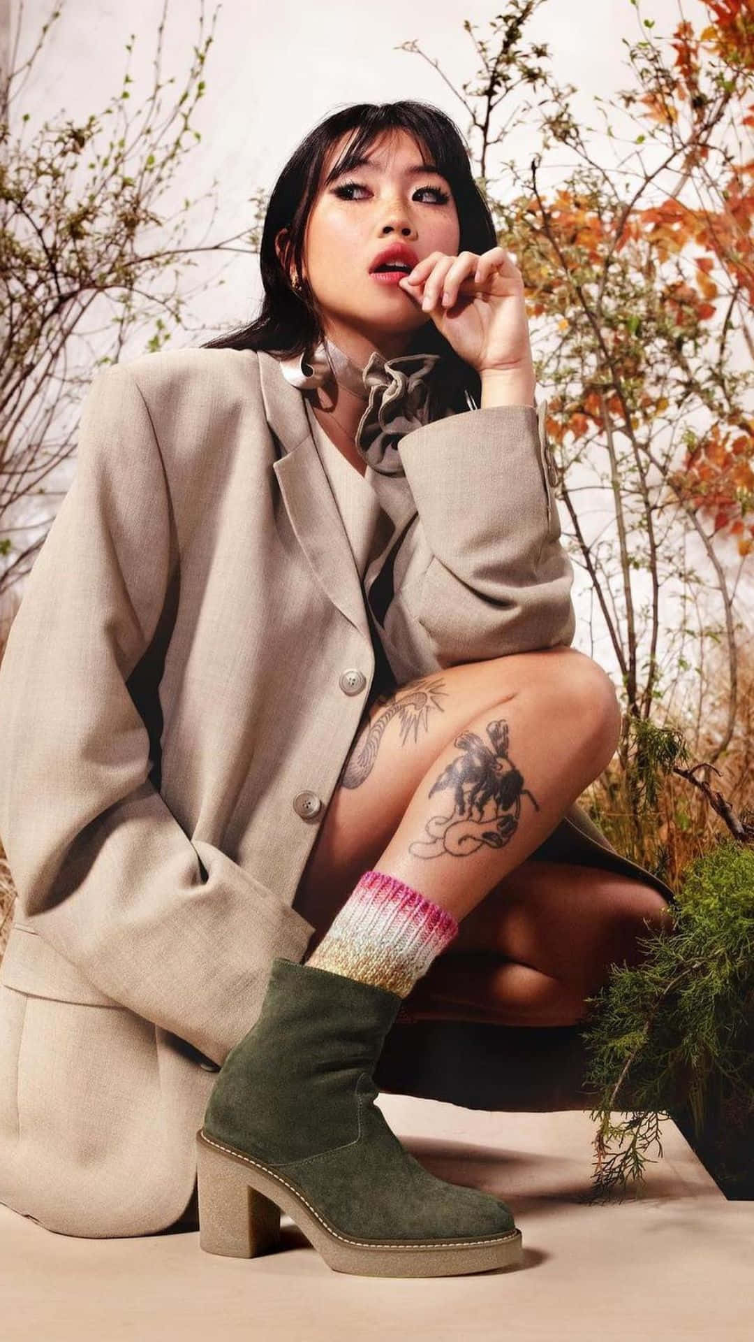  壁紙 — 一位時尚的紋身女性在戶外秋天的樹葉中蹲下，身穿米色西裝外套和綠色靴子。
