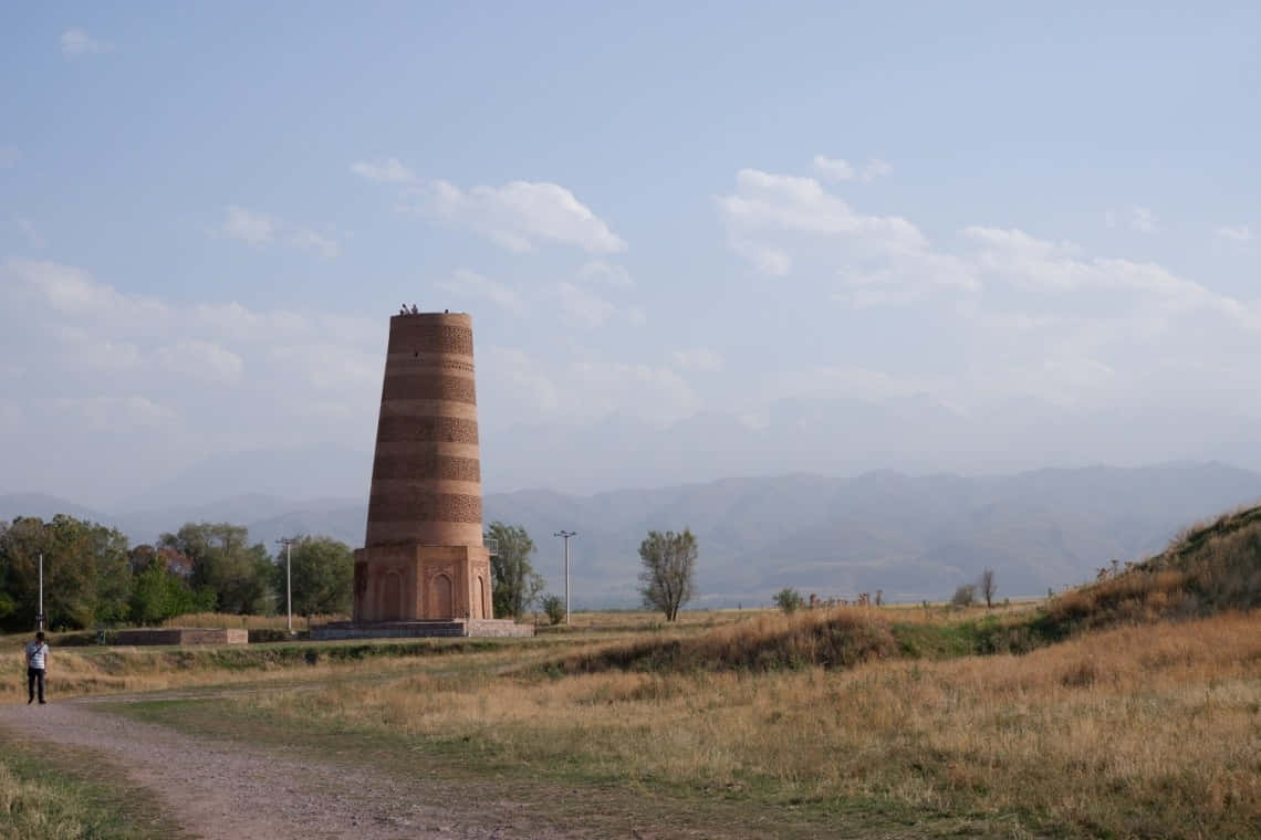 Kyrgyzstan 桌布 壁紙 — 一座高大的古老磚塔孤獨地矗立在乾燥的草地上，遠方有山脈，天空部分多雲。
