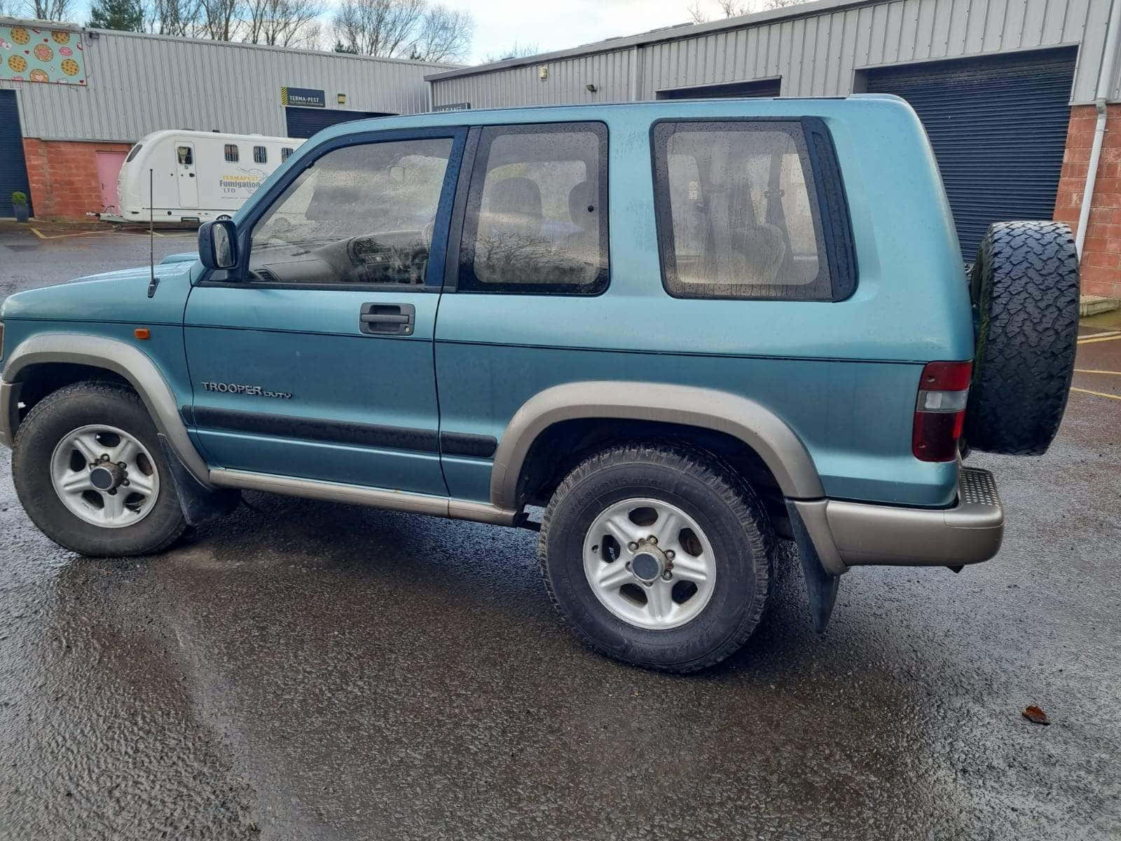  壁紙 — 一架青綠色鈴木Jimny Trooper SUV停泊在工業建築物前的濕路面上，天空多雲。