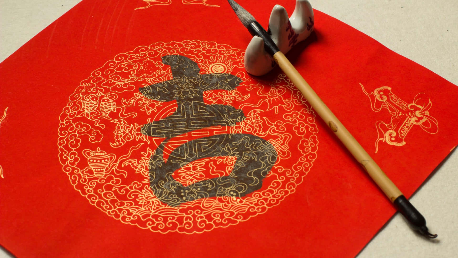  壁紙 — 一支傳統中國書法毛筆放在一張裝飾華麗的紅紙旁邊，紙上有一個被金色裝飾圖案包圍的大黑色「壽」字。