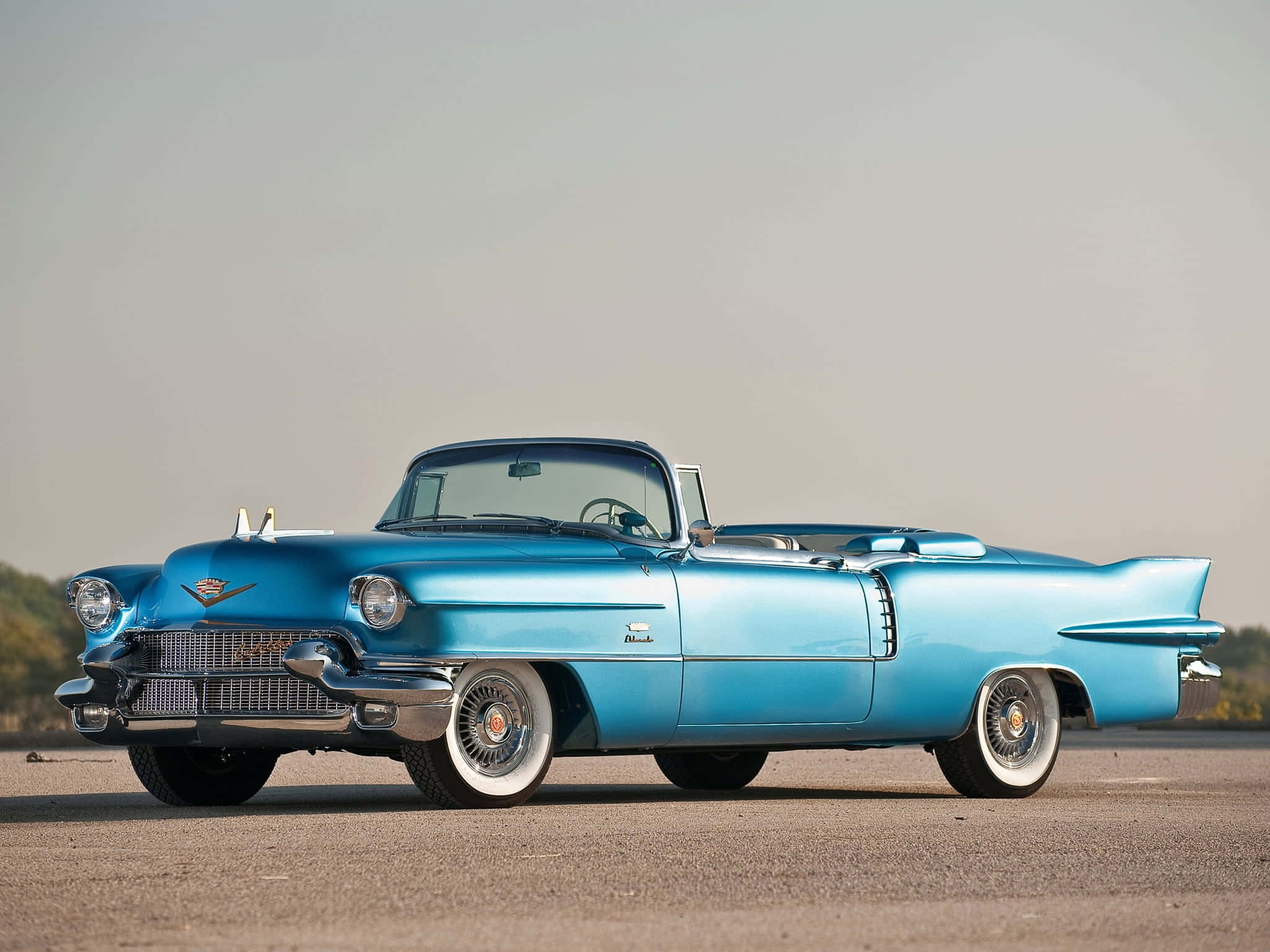 Cadillac Eldorado 桌布 壁紙 — 一輛鮮藍色嘅1959年Cadillac開篷車喺柔和日光下，喺一個有遠處樹木嘅開放鋪砌場地上閃閃發光。