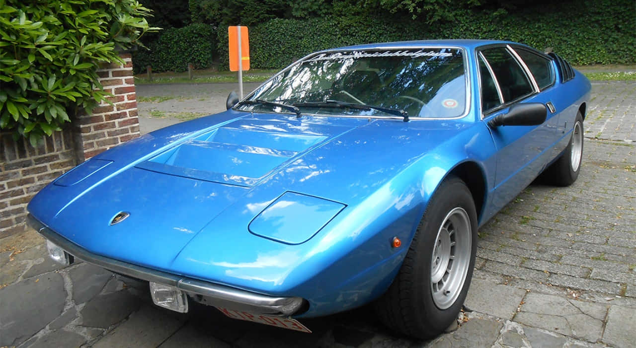 Lamborghini Urraco 桌布 壁紙 — 一架充滿活力的藍色經典林寶堅尼Countach停泊在石頭車道上，旁邊有綠植和磚牆。