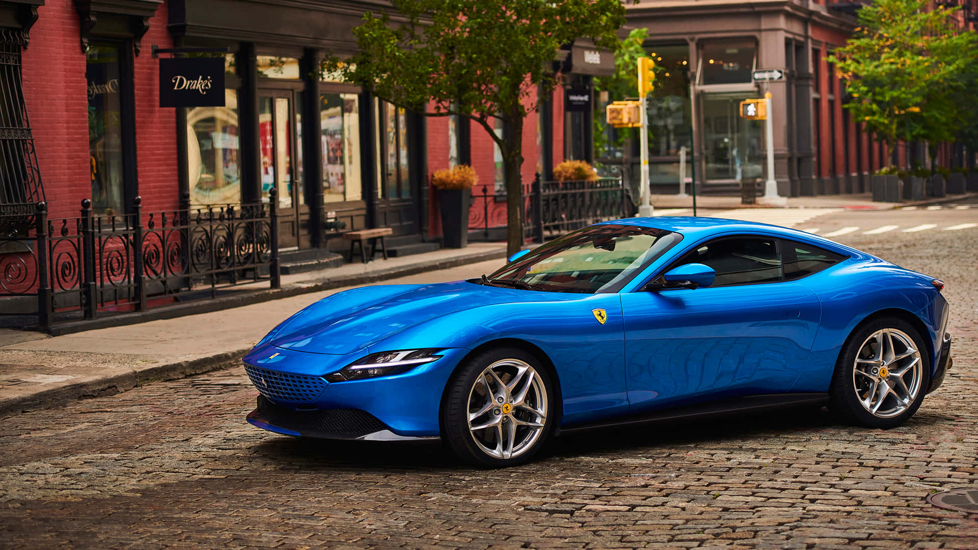 Ferrari Roma 桌布 壁紙 — 一輛充滿活力的藍色法拉利 Roma 停泊在鵝卵石街道上，兩旁是紅磚建築和綠樹。